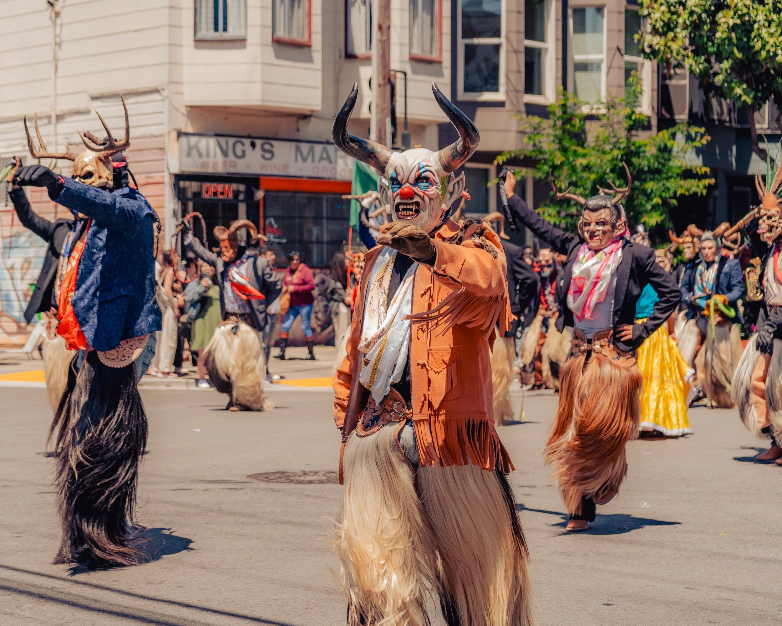 People in costumes participating in a parade, wearing masks with horns and fur, some dressed as mythical creatures or performers, marching on a city street with buildings and trees in the background.
