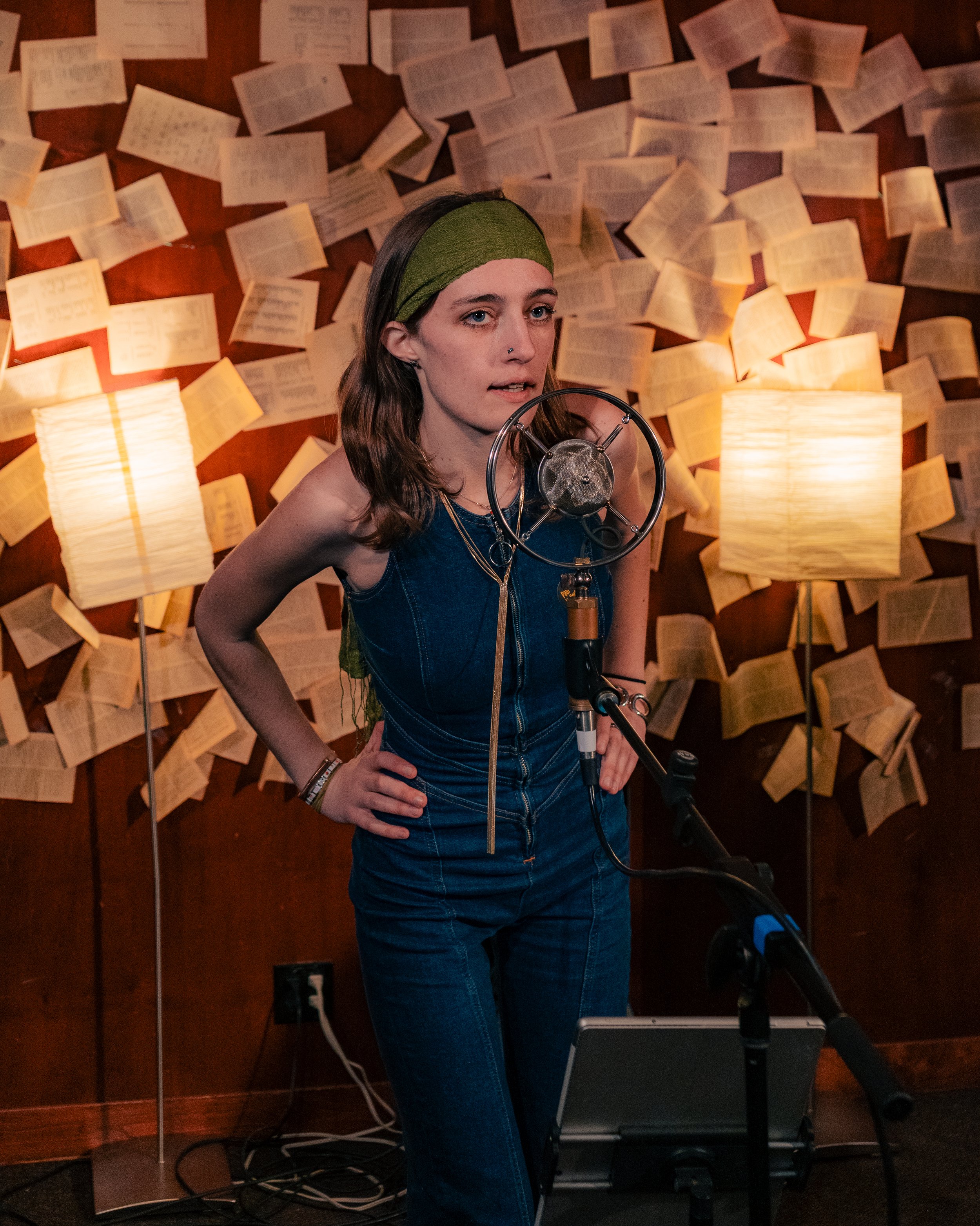 Young woman in denim jumpsuit singing into a vintage microphone in a recording studio with pages of sheet music or lyrics taped to the wallpaper behind her, illuminated by two warm table lamps.