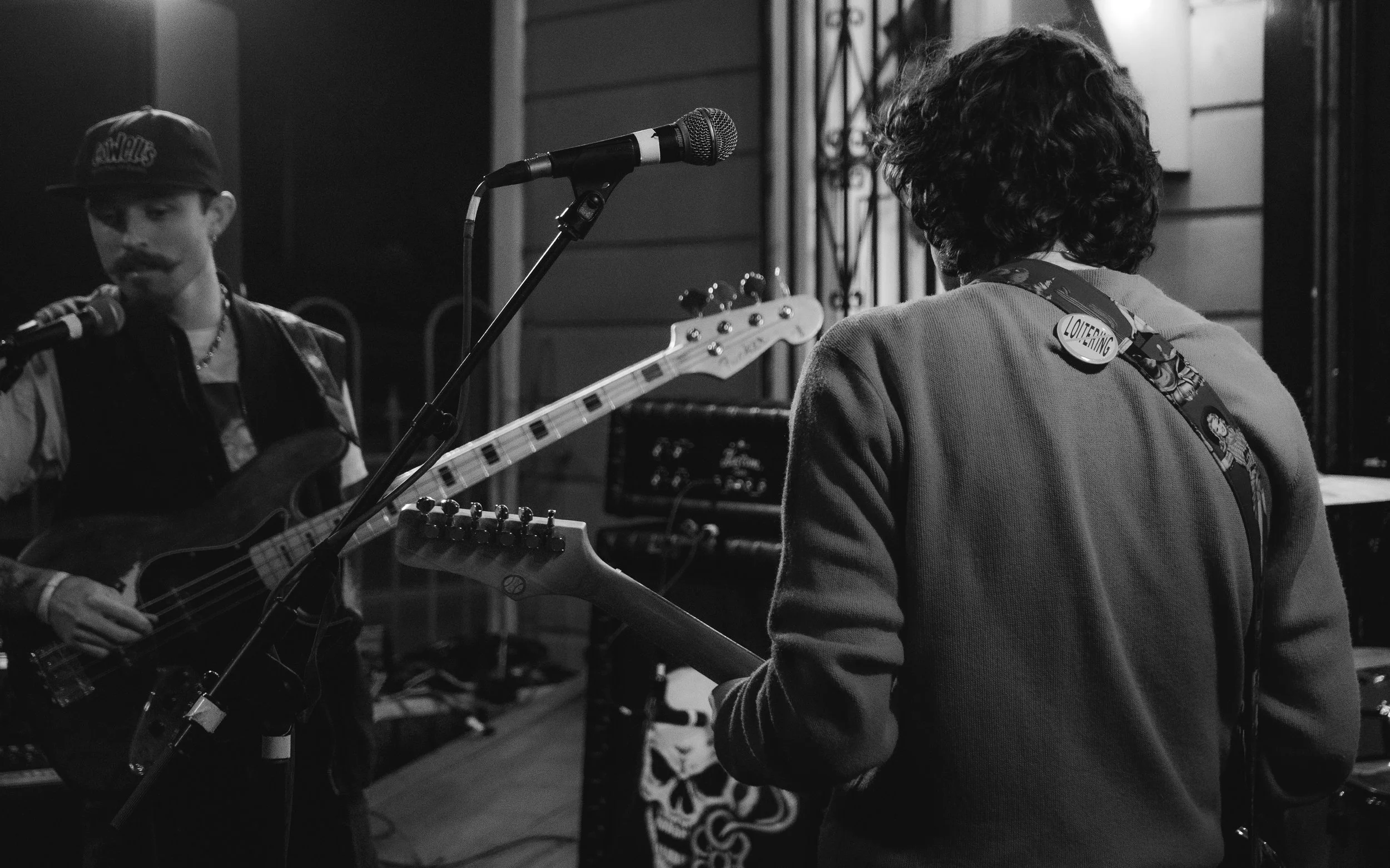 Two musicians practicing in a studio: one playing an electric bass guitar and singing into a microphone, the other playing an electric guitar with a patch reading 'LOITERING' on their strap. The scene is in black and white.
