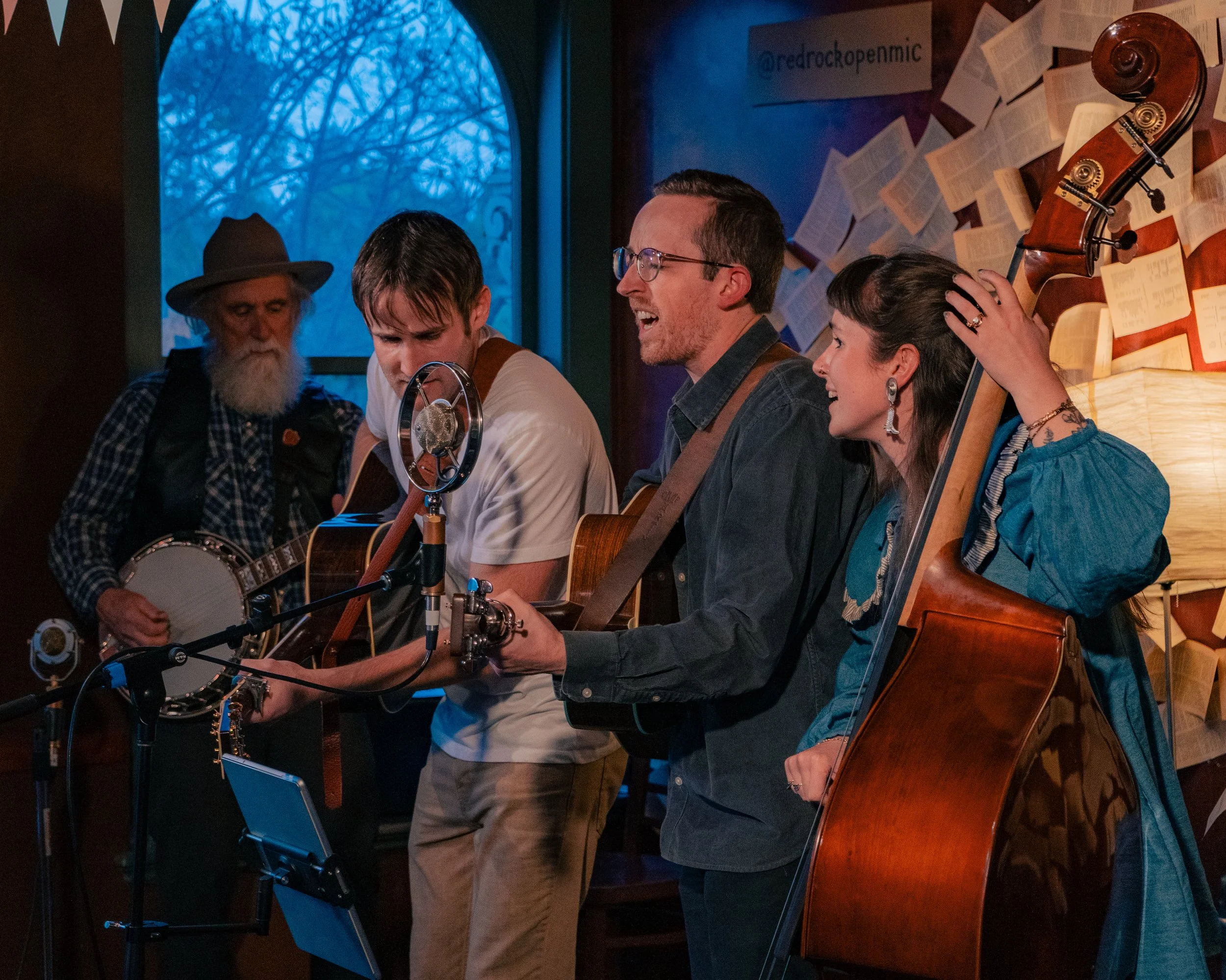 Four musicians performing together in a cozy, book-lined room with a window showing bare trees outside. They are playing guitars, a banjo, and a double bass, with music notes and papers on the wall behind them.