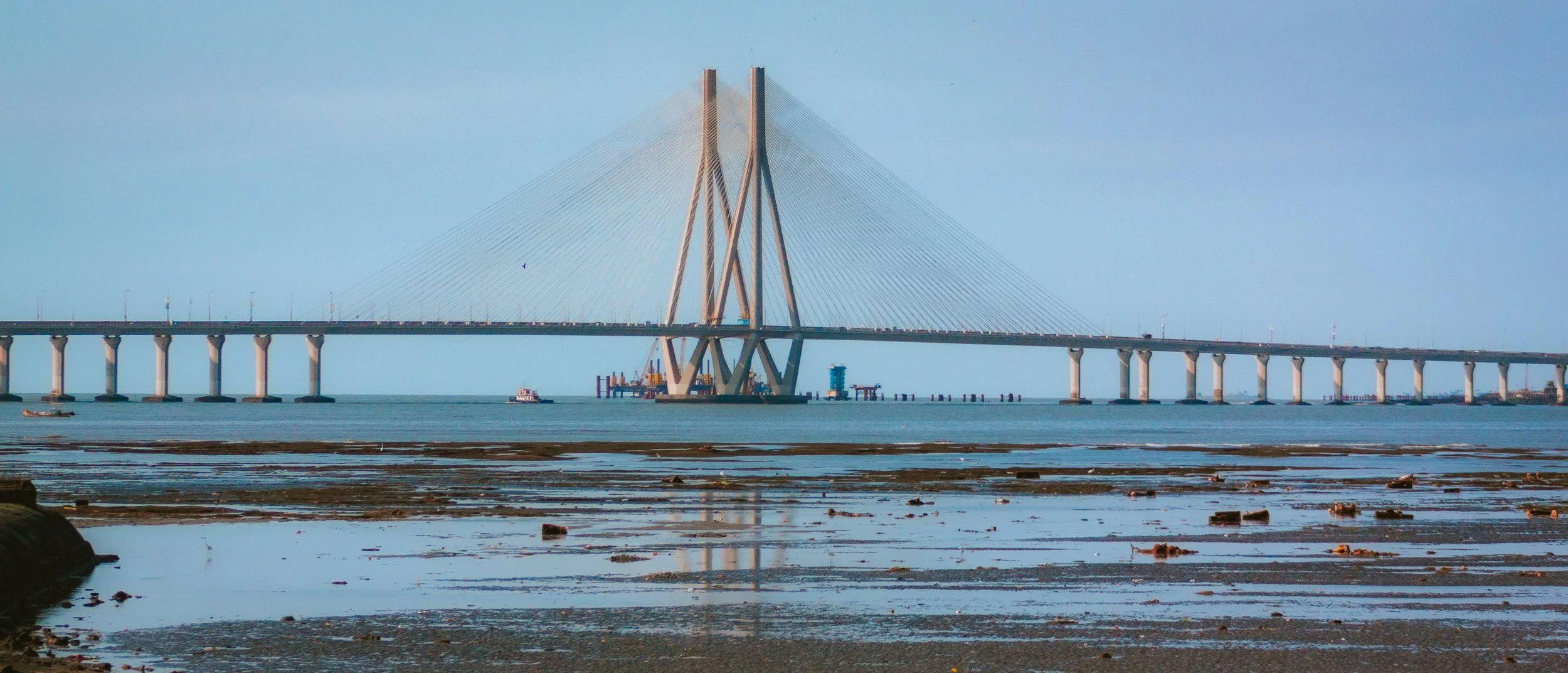 A large cable-stayed bridge extending across a body of water, with construction activity visible underneath, viewed from a low shoreline with wet sand and rocks in the foreground.