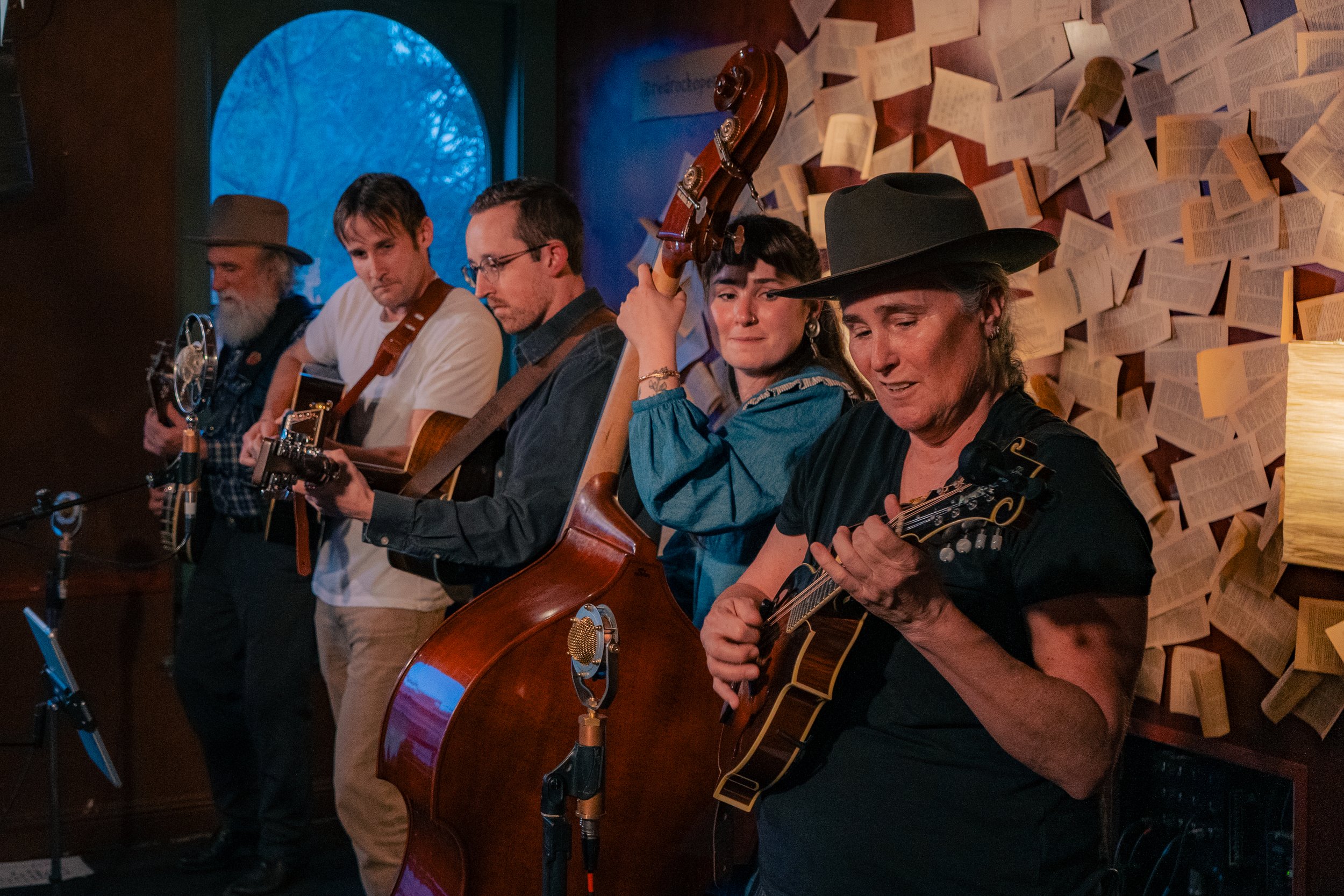 Five musicians performing in a cozy, dimly lit venue with a wall of open books behind them. They are playing various string instruments, including a guitar, bass, and mandolins, and appear emotionally engaged in their music.
