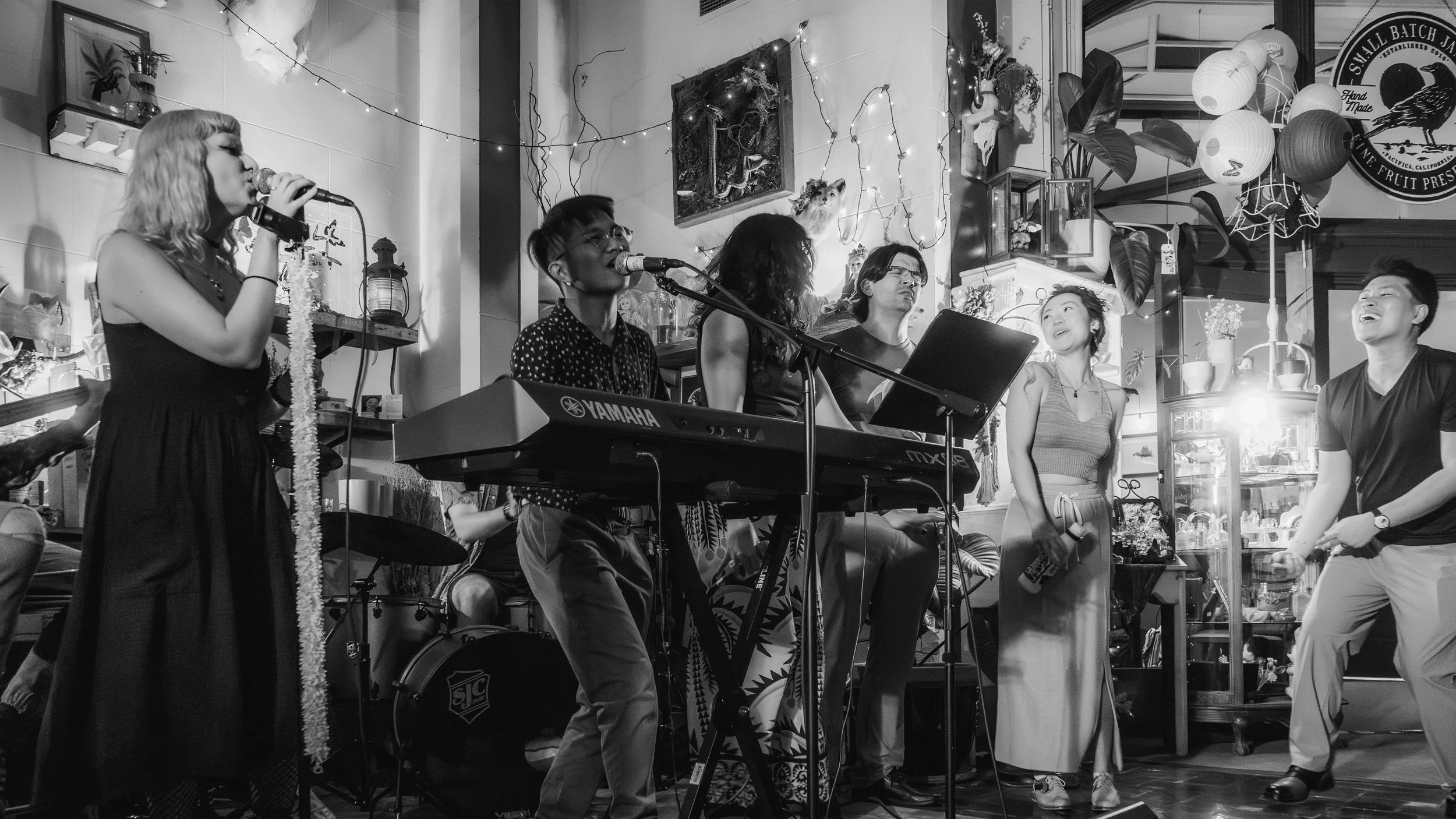 A group of people performing music and singing at a cozy indoor venue decorated with string lights, plants, and paper lanterns.