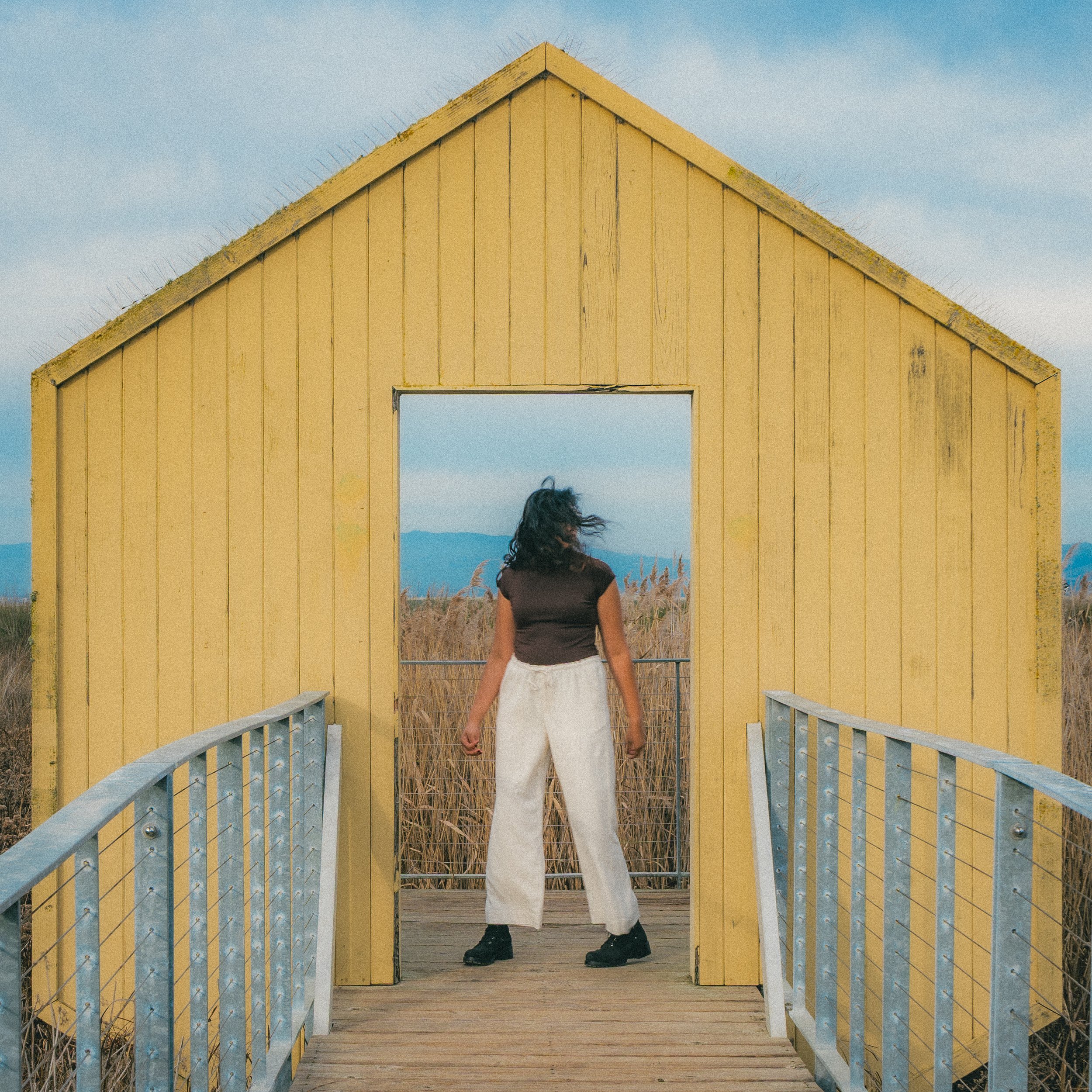 Woman standing at the entrance of a small yellow wooden house with a triangular roof, overlooking a field of tall grass with mountains in the distance.