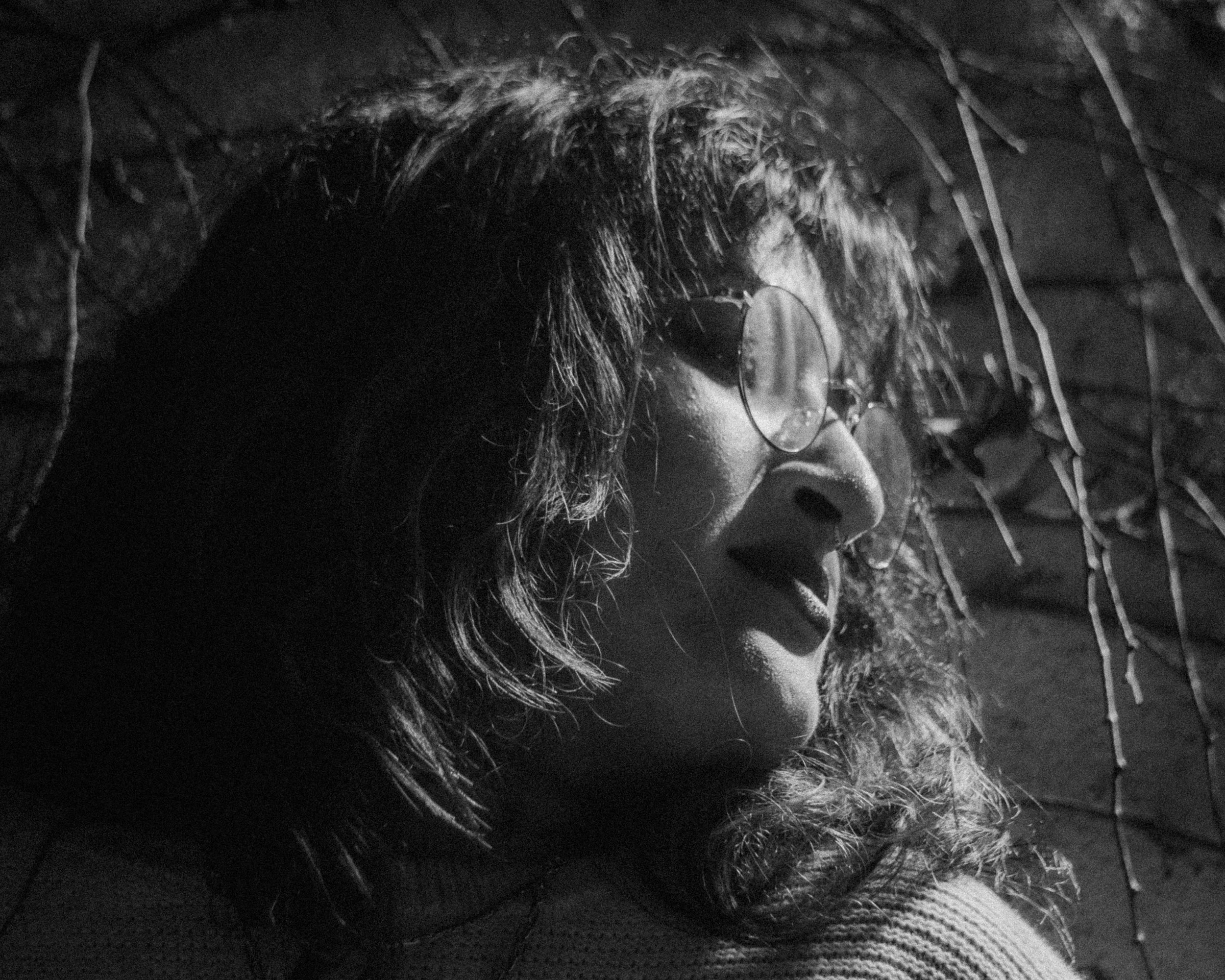 A black and white photo of a woman with glasses and curly hair looking to the side, outdoors with dried plants or branches around her.
