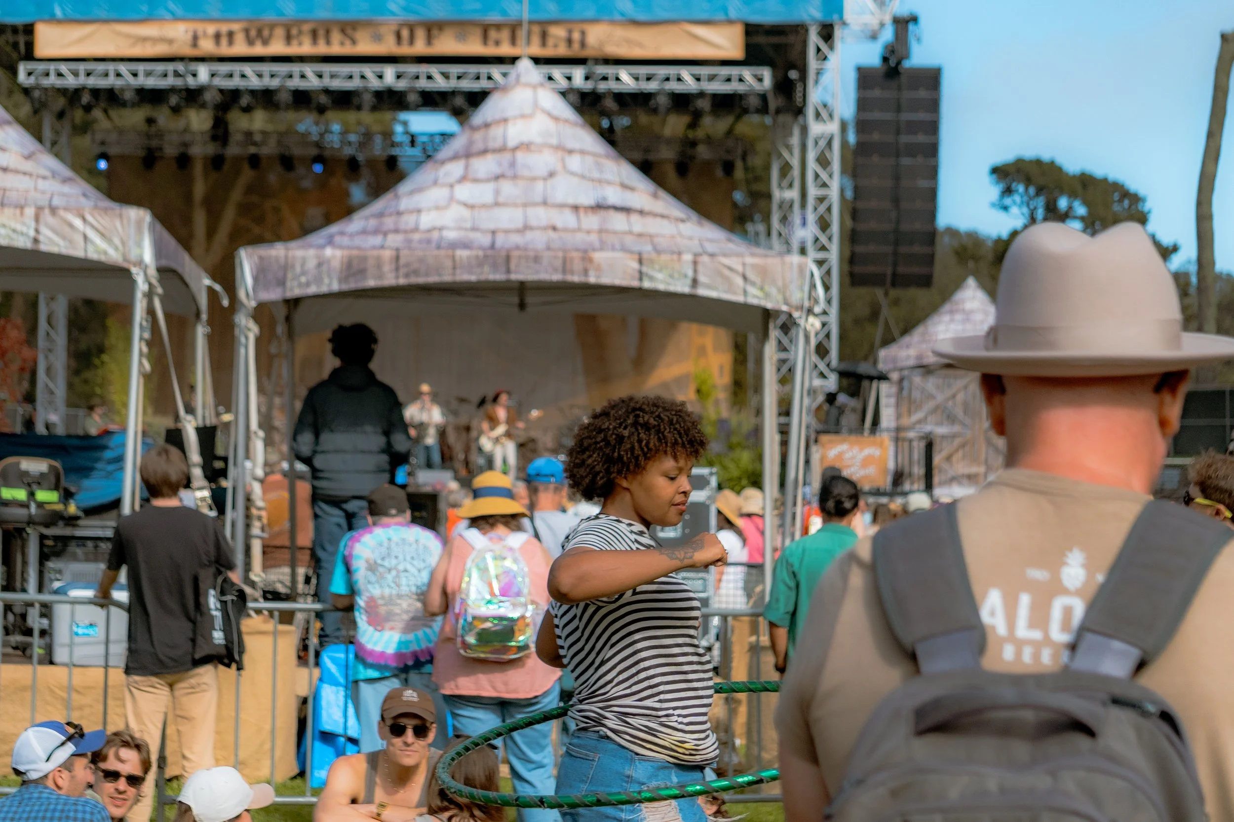 People attending an outdoor music festival with a stage and band performing in the background. Many individuals are wearing hats and backpacks, some are taking photos or looking at the stage.