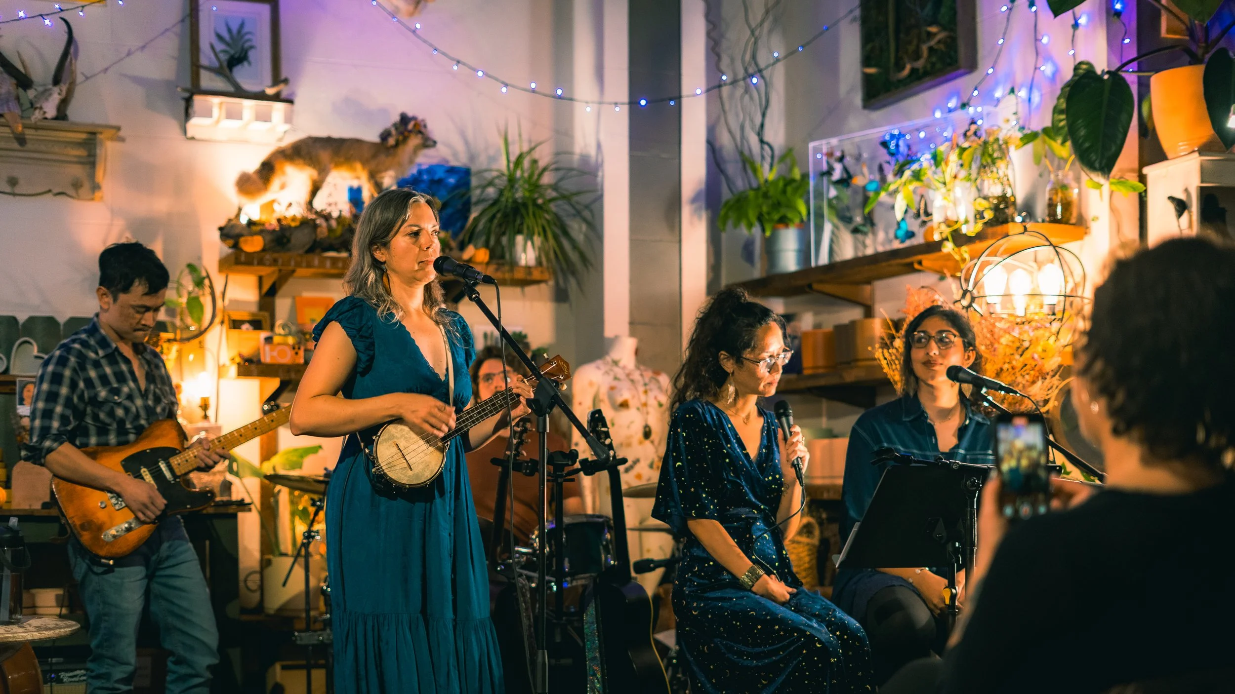 A band performing live music indoors, with three women singing into microphones and a man playing guitar, surrounded by decorative plants and string lights.