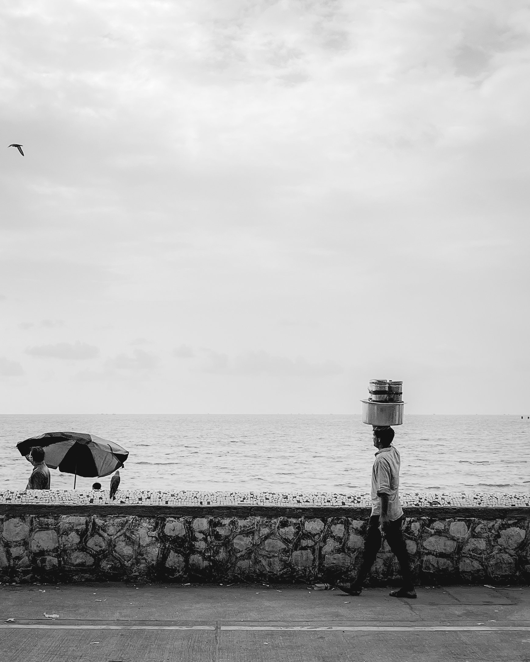 A man walking along a seafront promenade with a stone boundary, carrying two large containers on his head. In the background, a woman with a wide-brimmed umbrella is walking by the ocean, and a bird flying in the sky is visible.