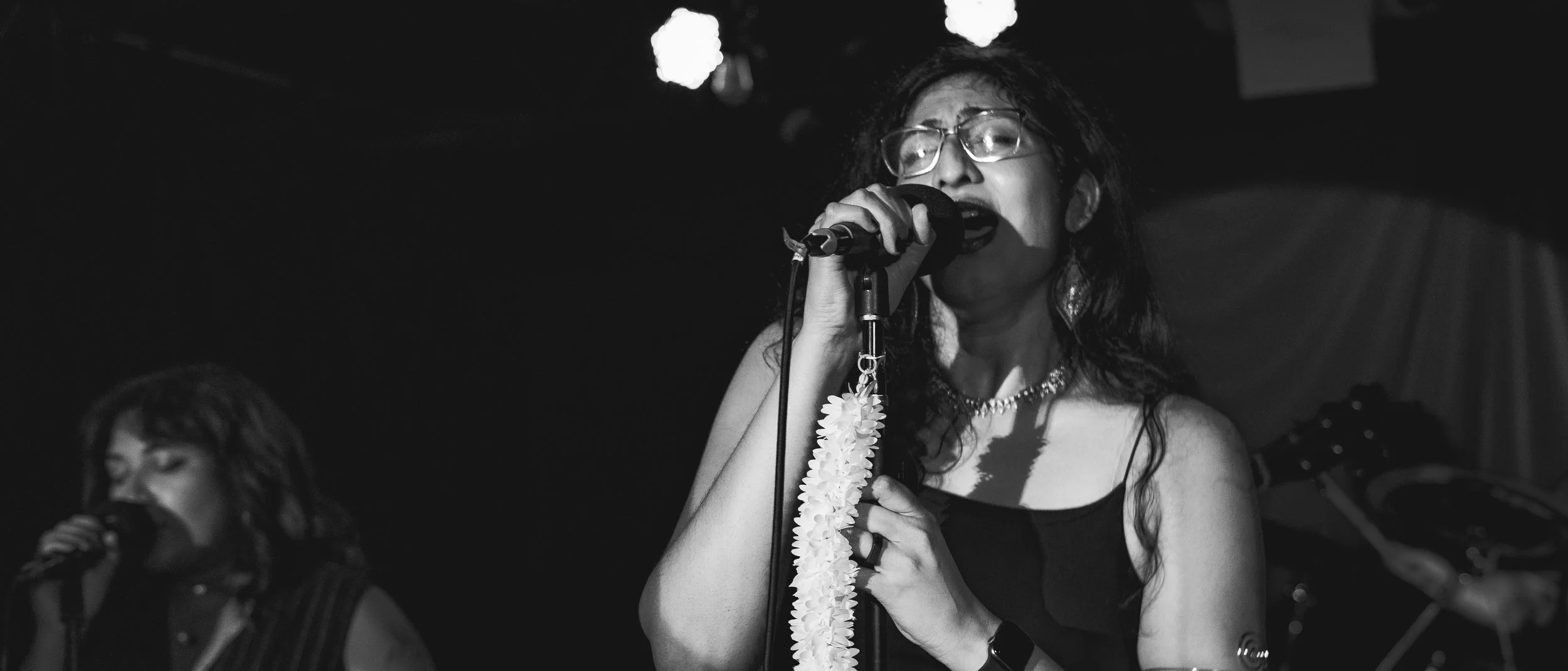 Black and white photo of a woman singing into a microphone on stage, with another woman singing in the background, surrounded by stage lights.