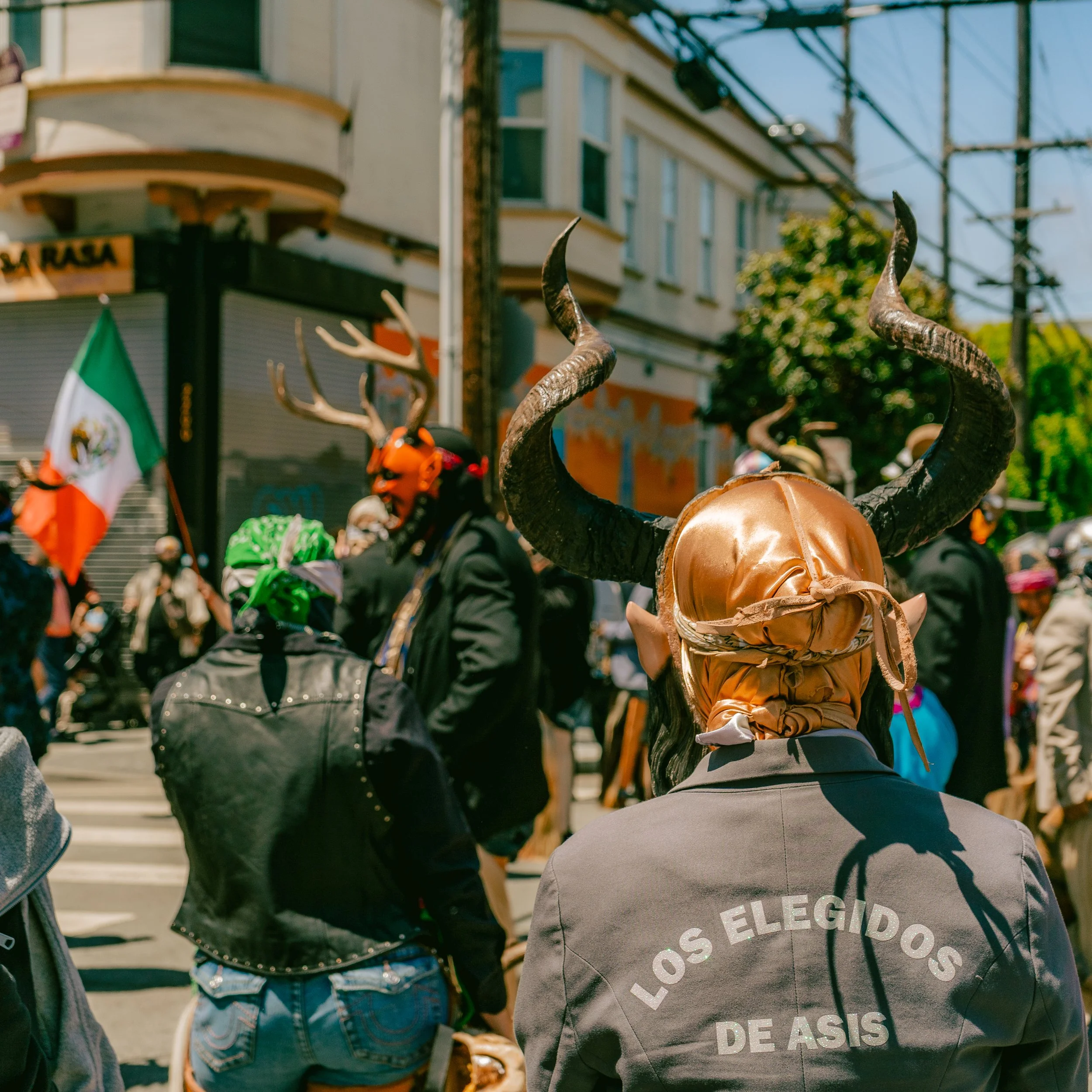 People in a street protest wearing costumes with horns and masks, with one person wearing a costume and a jacket that reads 'LOS ELEGIDOS DE AXSIS', and others holding a Mexican flag.