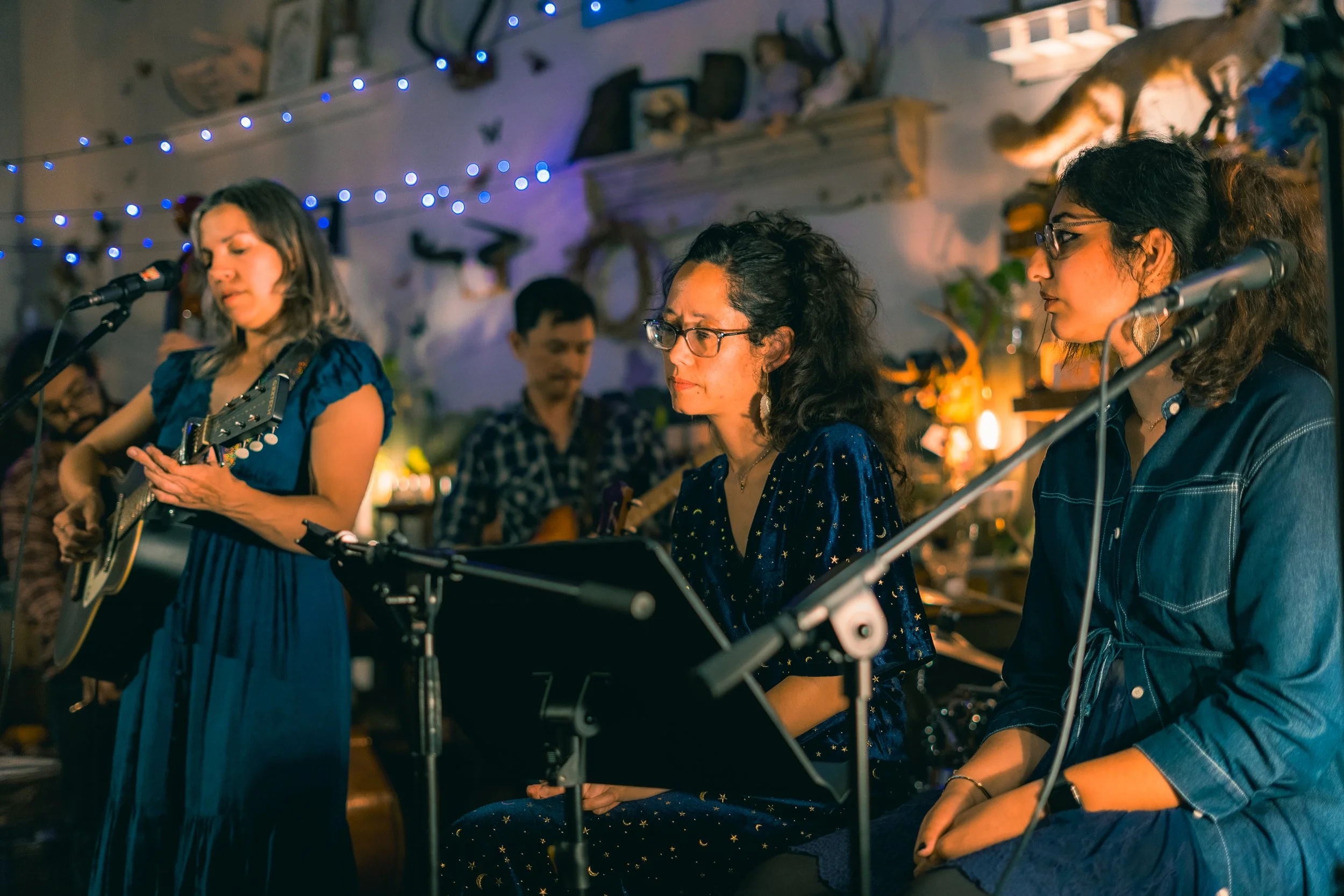Group of women performing music indoors, one playing guitar, others singing with microphones, decorated with fairy lights and plants.
