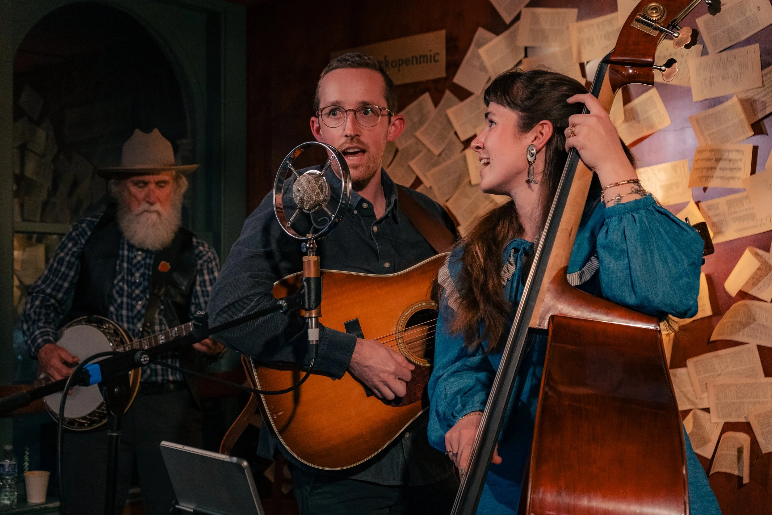 Three musicians performing in a recording studio: a man playing an acoustic guitar singing into a microphone, a woman playing a double bass, and an older man with a beard playing a banjo in the background.