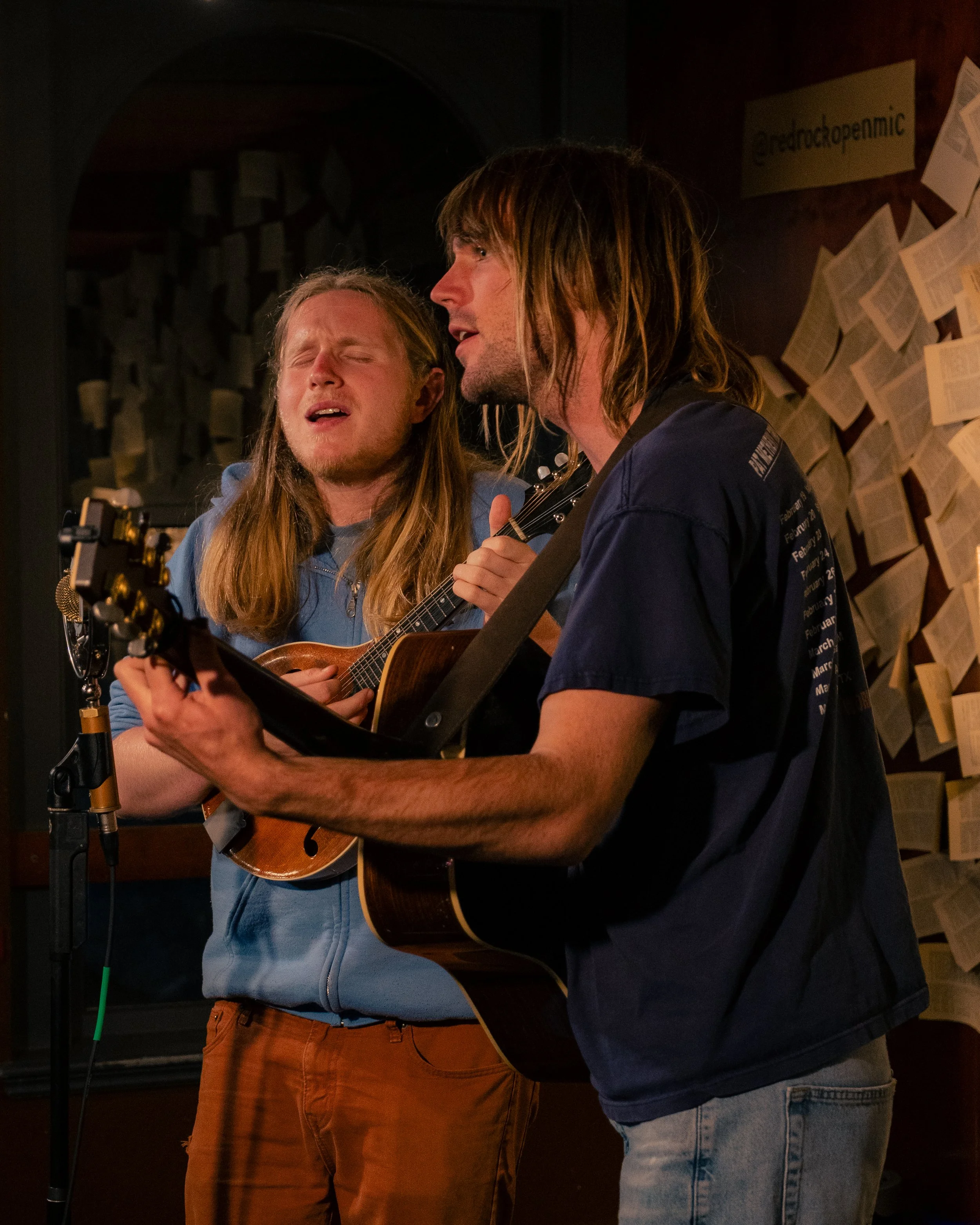 Two men with long hair singing and playing guitars in a dimly lit room, with pages of sheet music on the wall behind them.