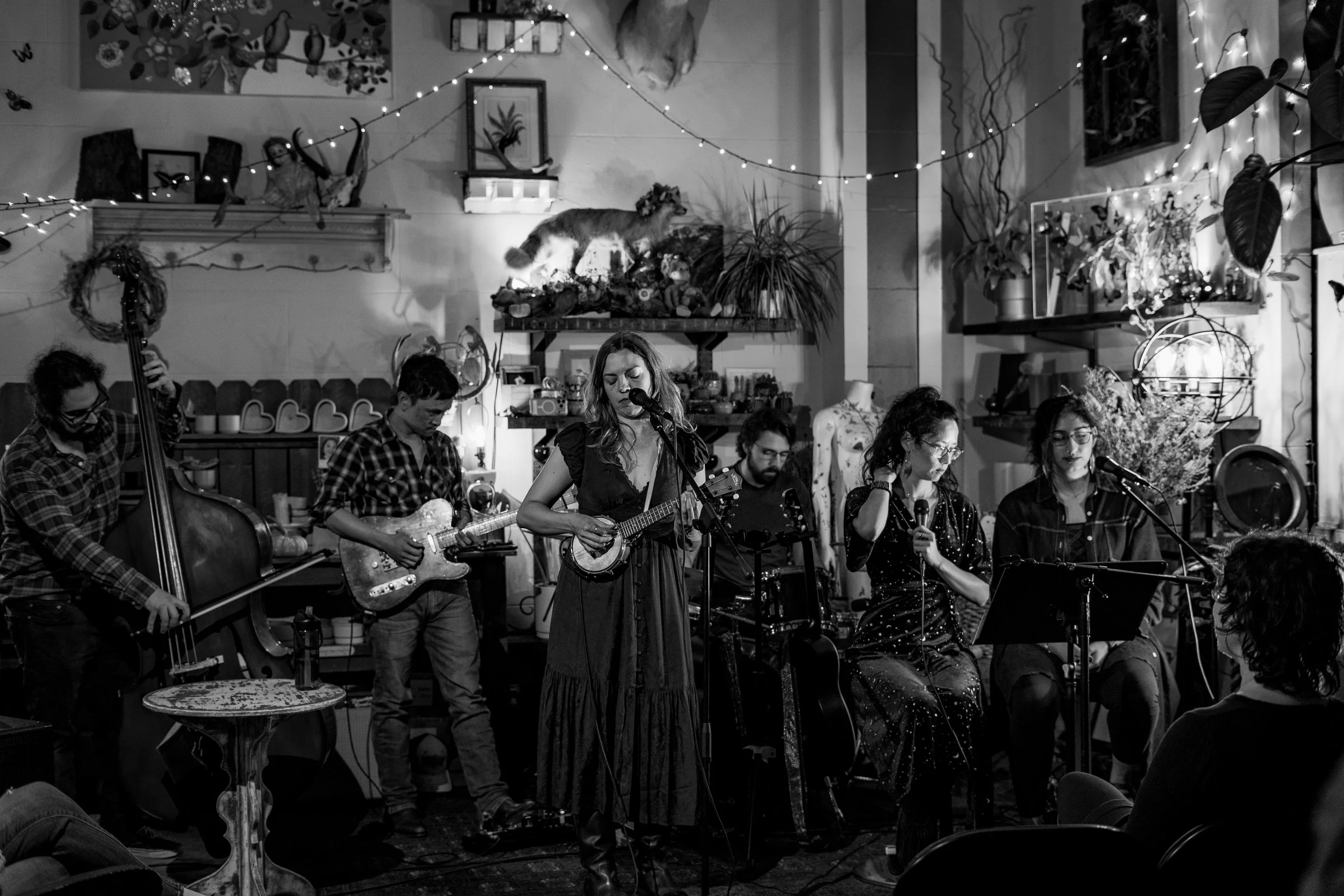 A band performing with four women singing and playing instruments, surrounded by Christmas decorations in an intimate indoor setting.