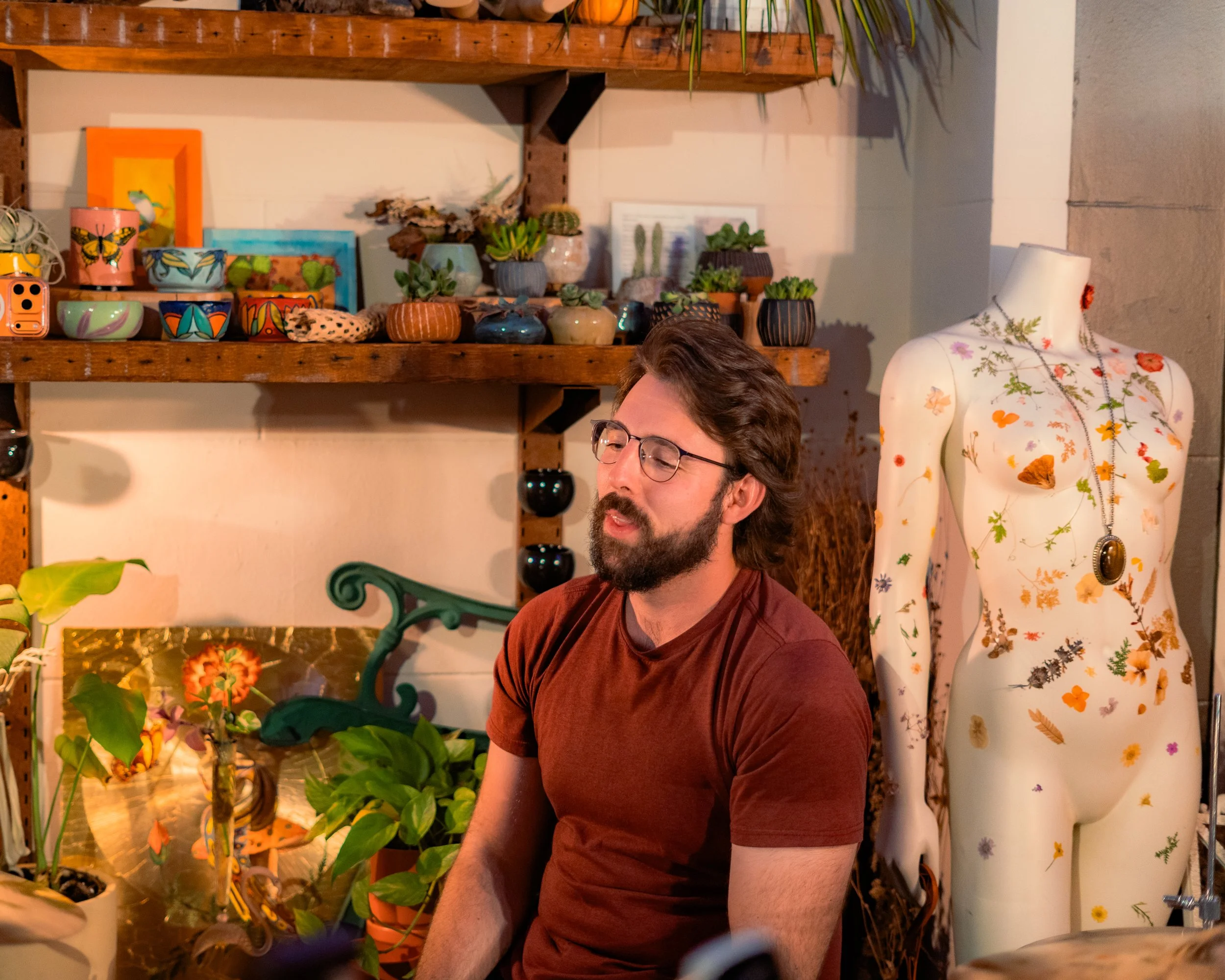 A man with glasses and a beard wearing a maroon t-shirt is sitting indoors surrounded by plants, pottery, and a floral mannequin adorned with necklaces. The background features wooden shelves with small potted plants and decorative items.