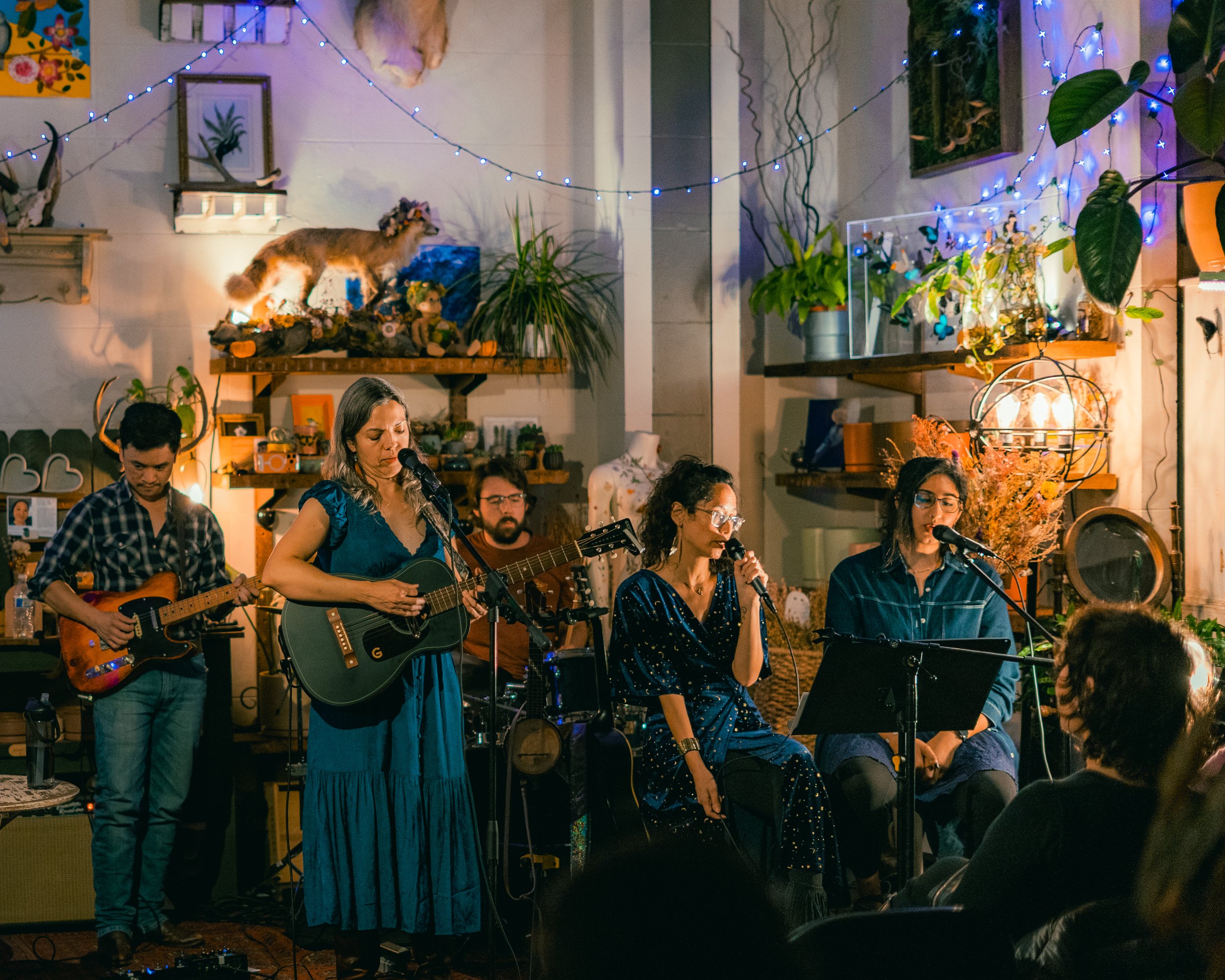 A live music performance indoors featuring five musicians. Two women and one man are singing into microphones, with one woman playing an acoustic guitar. Two men are playing electric guitars in the background. The setting has decorative plants, strin