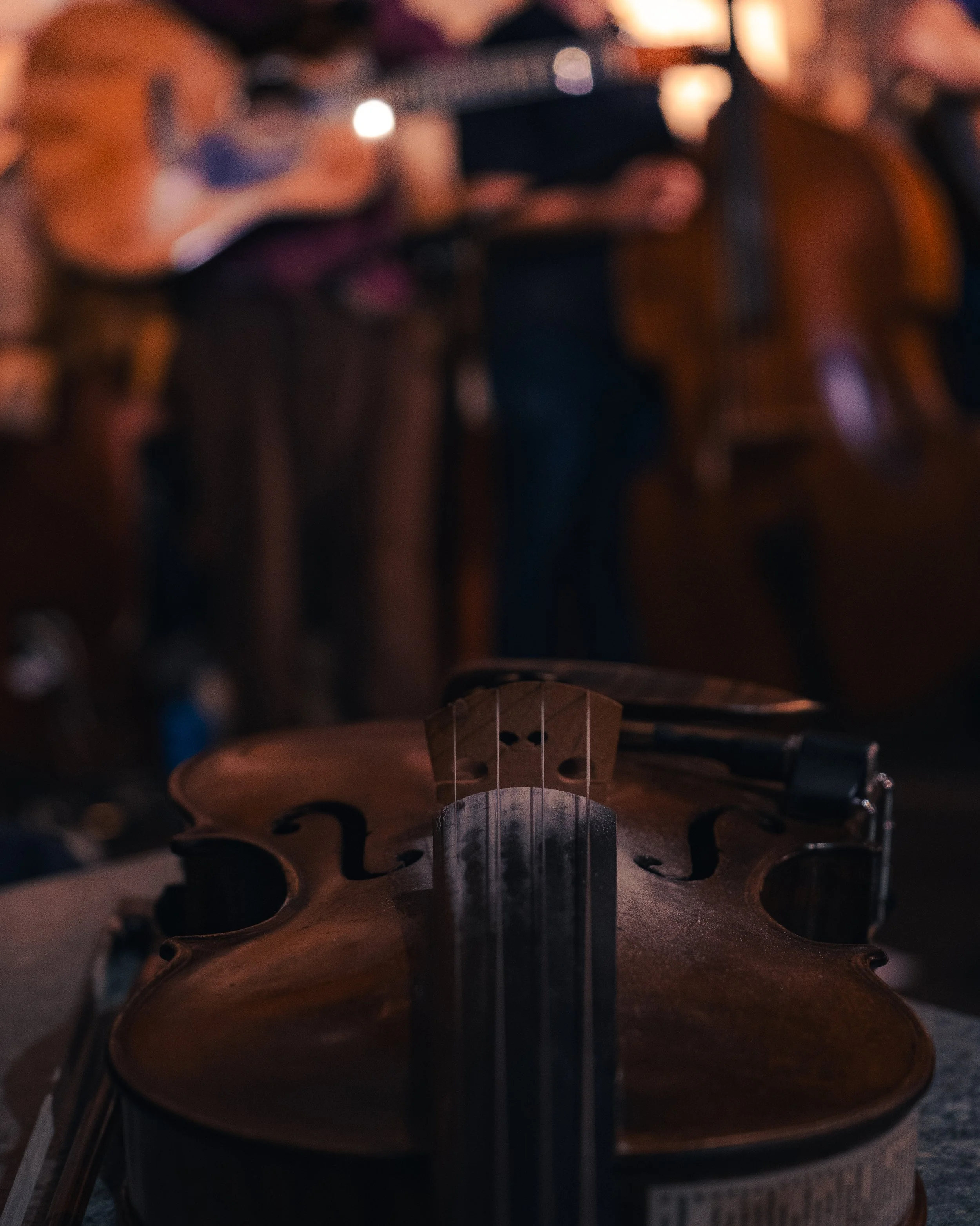 Close-up of a violin on a table with musicians playing guitars in the blurred background at night.