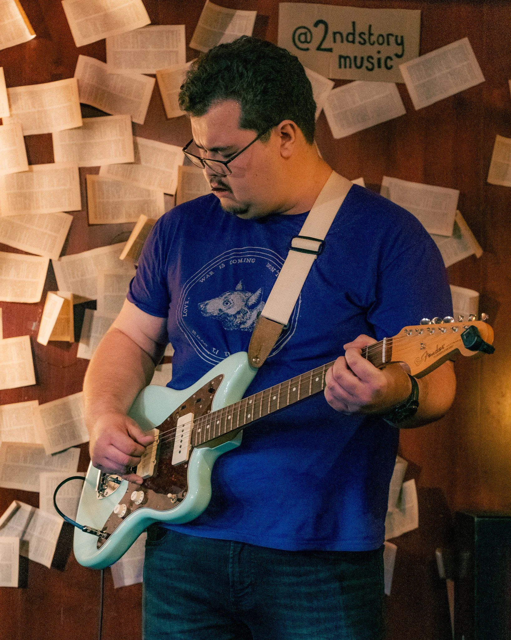 A man wearing glasses and a blue t-shirt playing an electric guitar in a room decorated with open books on the wall and a sign that reads '@2ndstory music'.