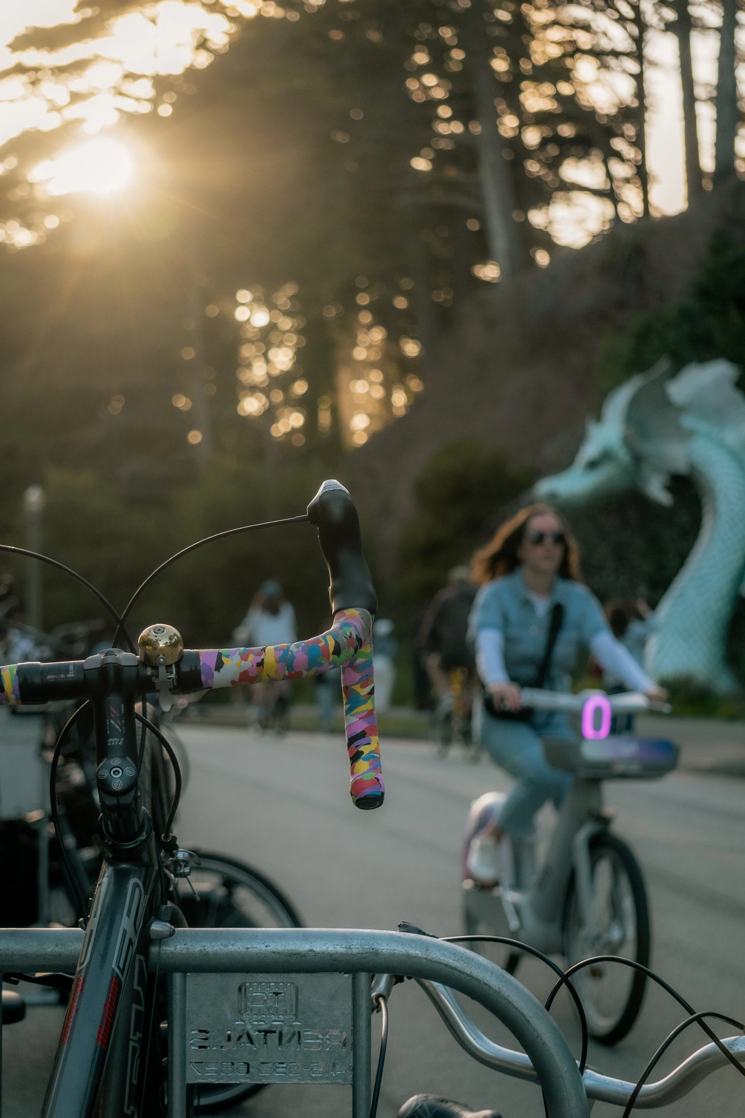 Close-up of a bicycle handlebar with a colorful grip in the foreground, and a woman riding a bike with a dragon sculpture in the background during sunset.