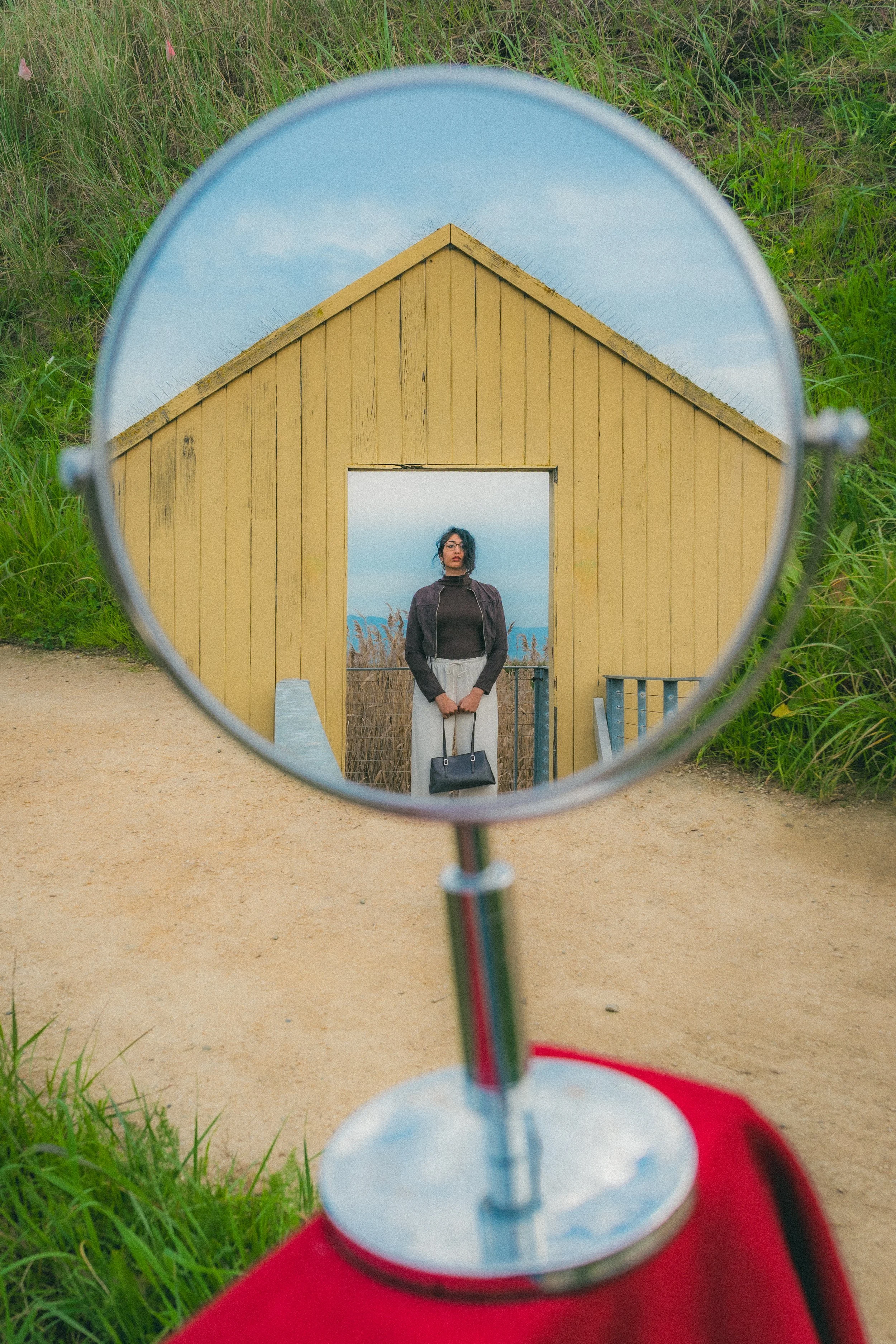 A person standing in front of a yellow wooden structure, viewed through a round mirror mounted on a red surface, with grass and a cloudy sky in the background.