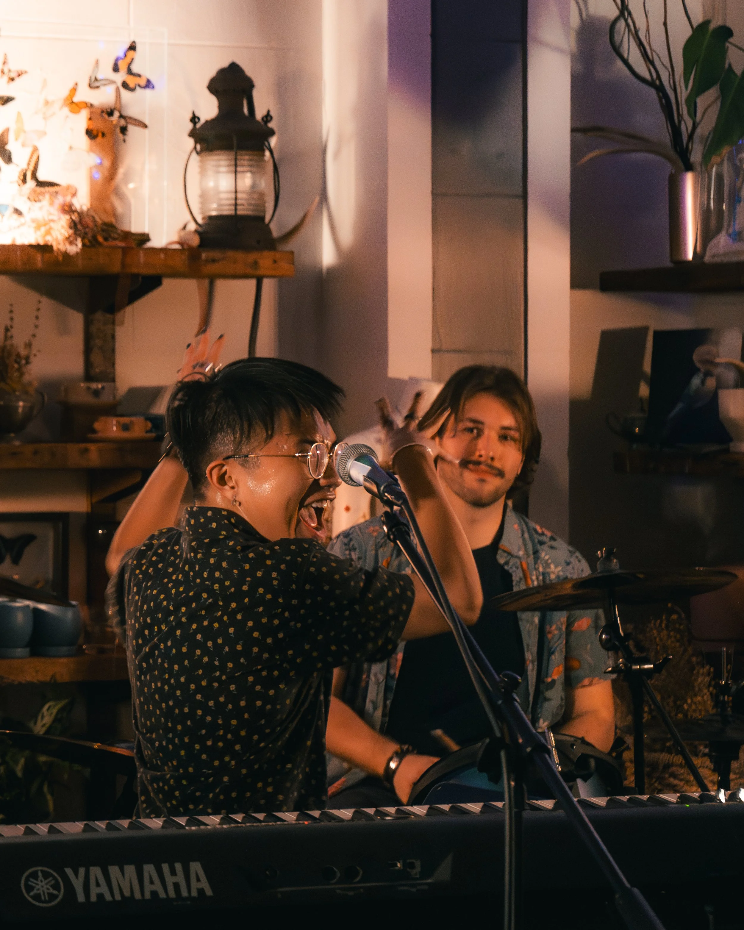A live performance scene with a woman singing into a microphone and playing a keyboard on stage, accompanied by a man playing drums in a cozy indoor setting decorated with plants, lanterns, and butterfly wall art.