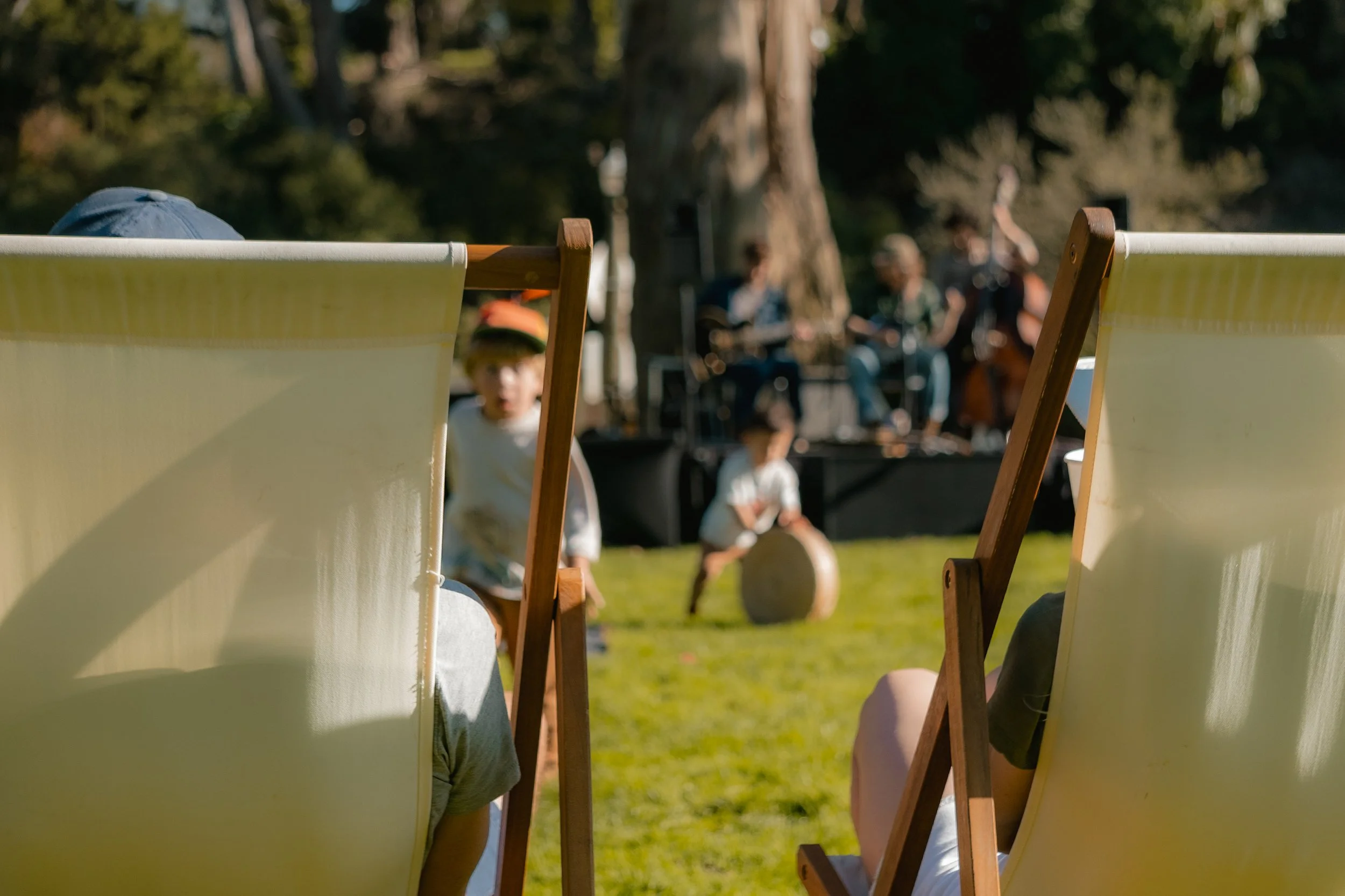 Magical outdoor scene with children in summer, with a band playing music in the background, viewers are on shaded deck chairs.