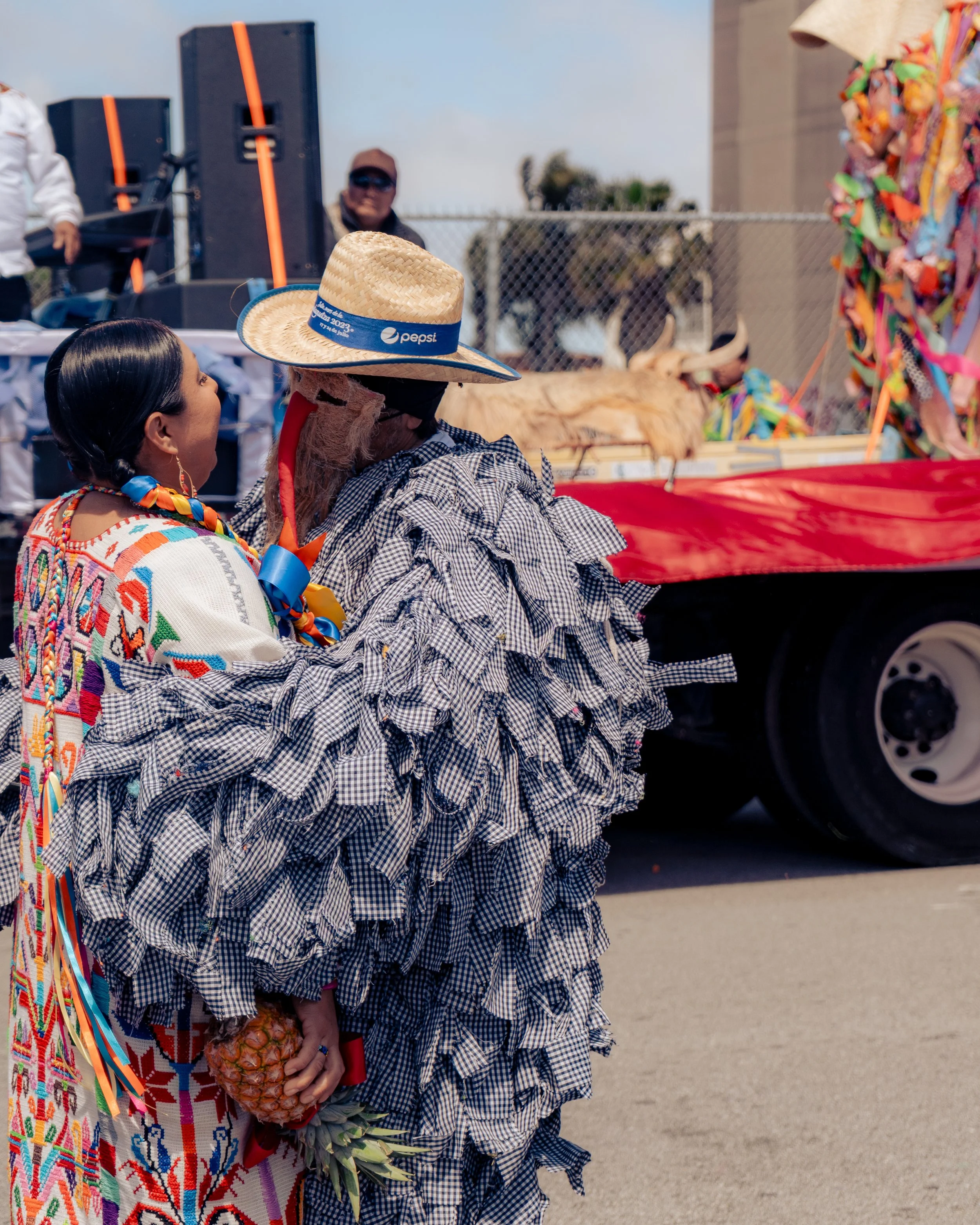 People dressed in traditional Mexican clothing participating in a parade, with one holding a pineapple. A decorated float with a cow is visible in the background.