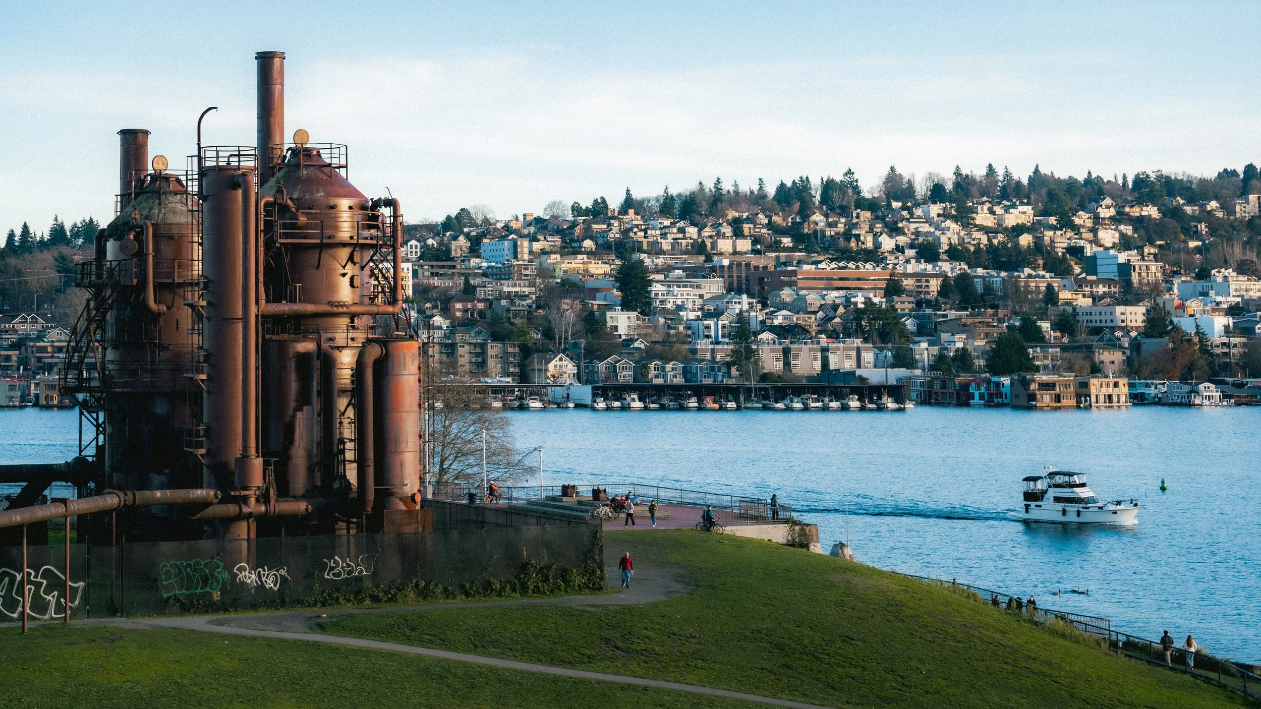 A waterfront scene with a rusty industrial structure to the left, a green grassy area with people walking and biking, a small white boat on the water, and a hillside filled with residential houses in the background.