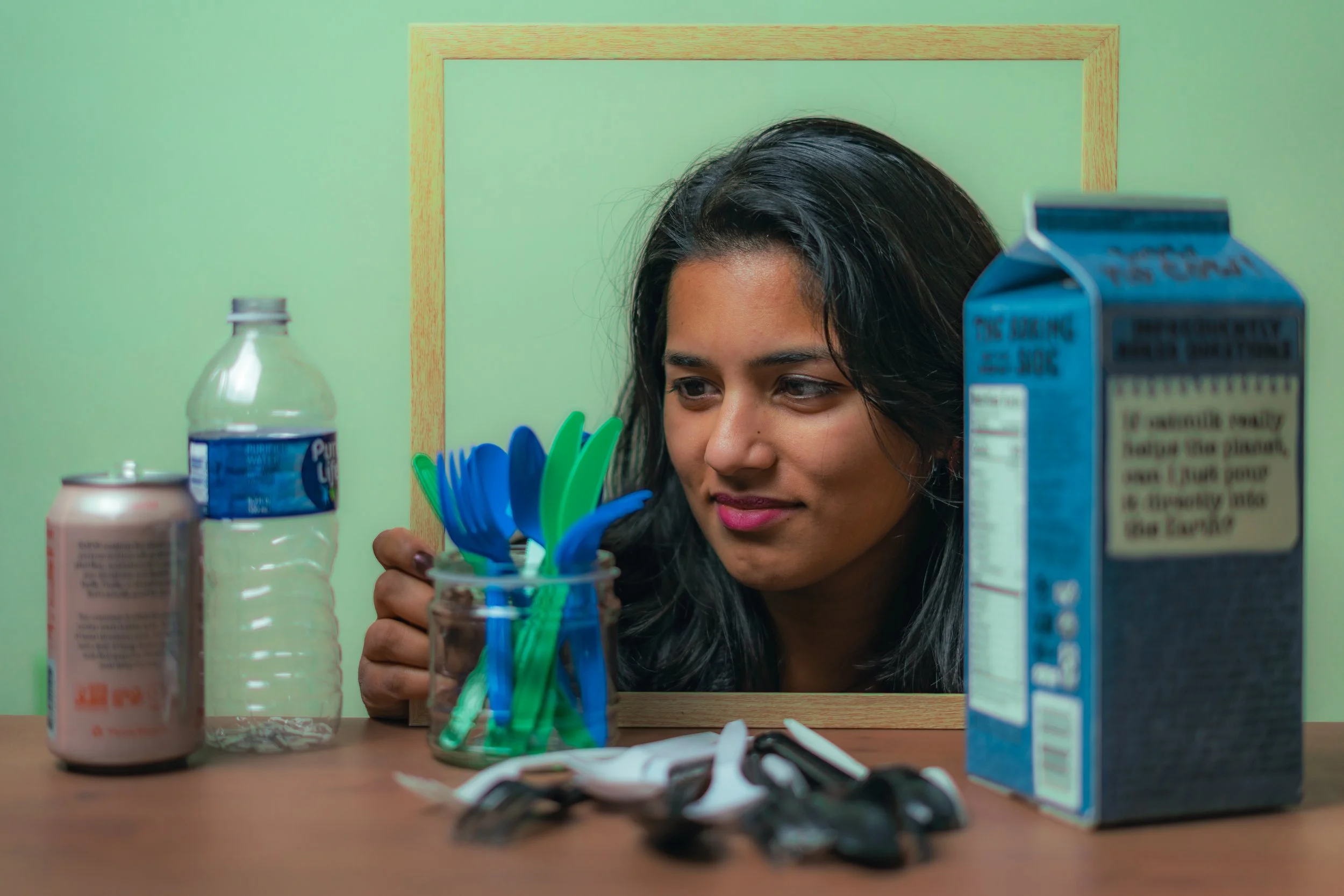 A woman looks at a mirror on a table with various items, including a water bottle, a soda can, a juice box, sunglasses, and a container of colorful spoons.