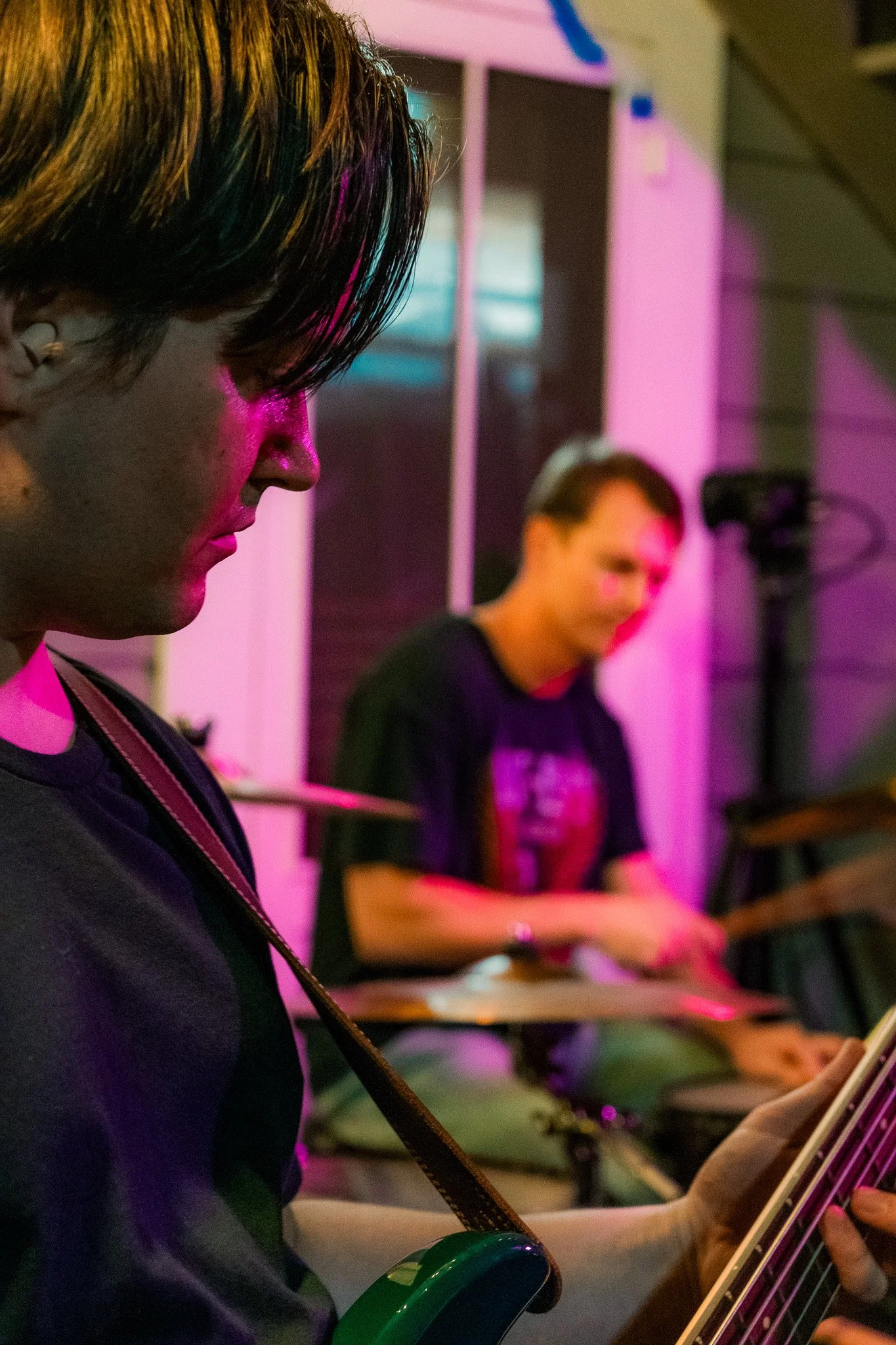A person playing guitar with another person playing drums in the background, illuminated by pink and purple lighting.