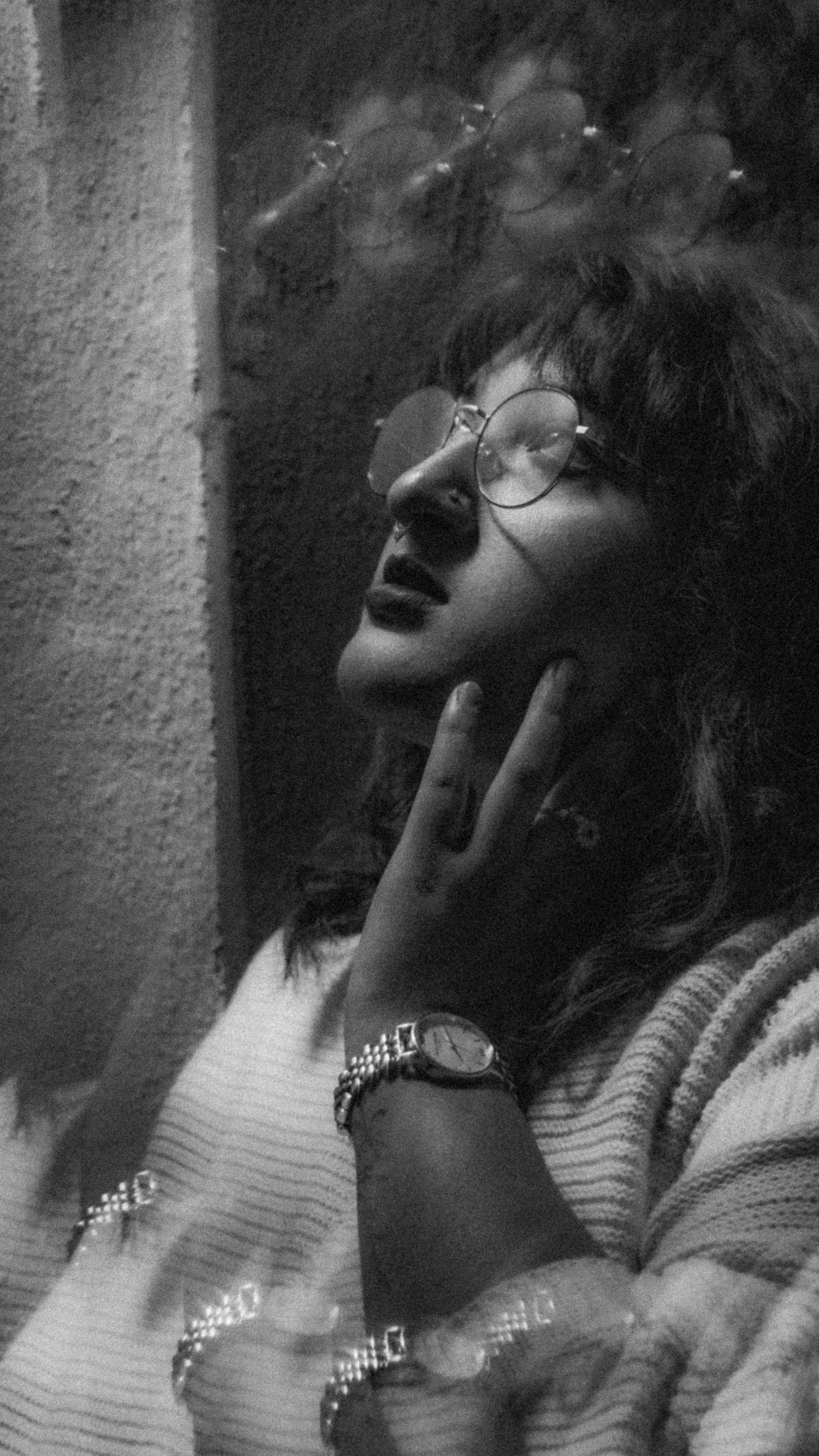 A black and white photo of a woman with glasses, dark lipstick, and wavy hair, touching her neck with her hand, wearing jewelry including a watch and bracelets, against a textured wall.