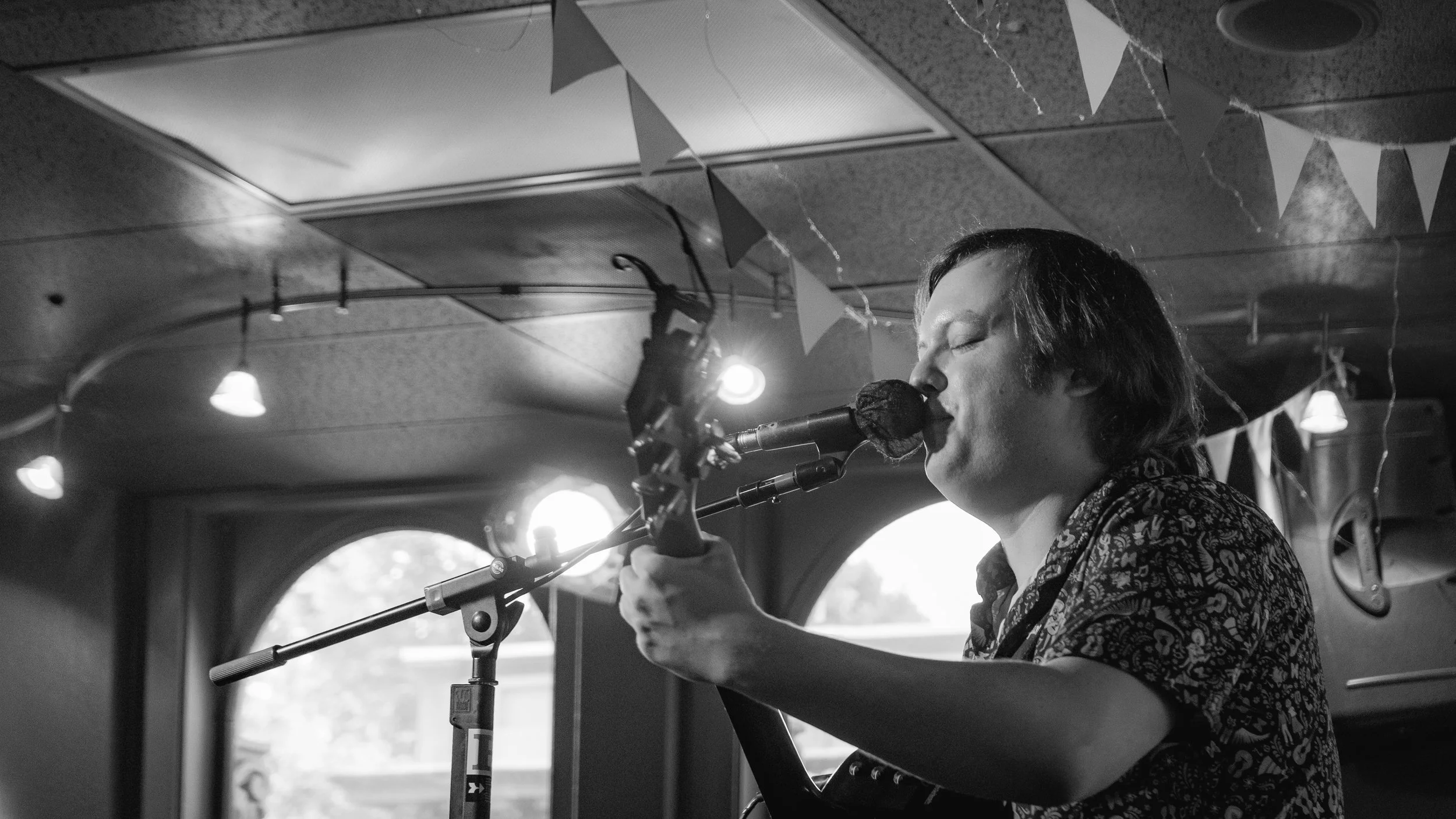 A man with medium-length hair singing and playing guitar in a dimly lit room decorated with small flags and string lights, with sunlight coming through a window in the background.