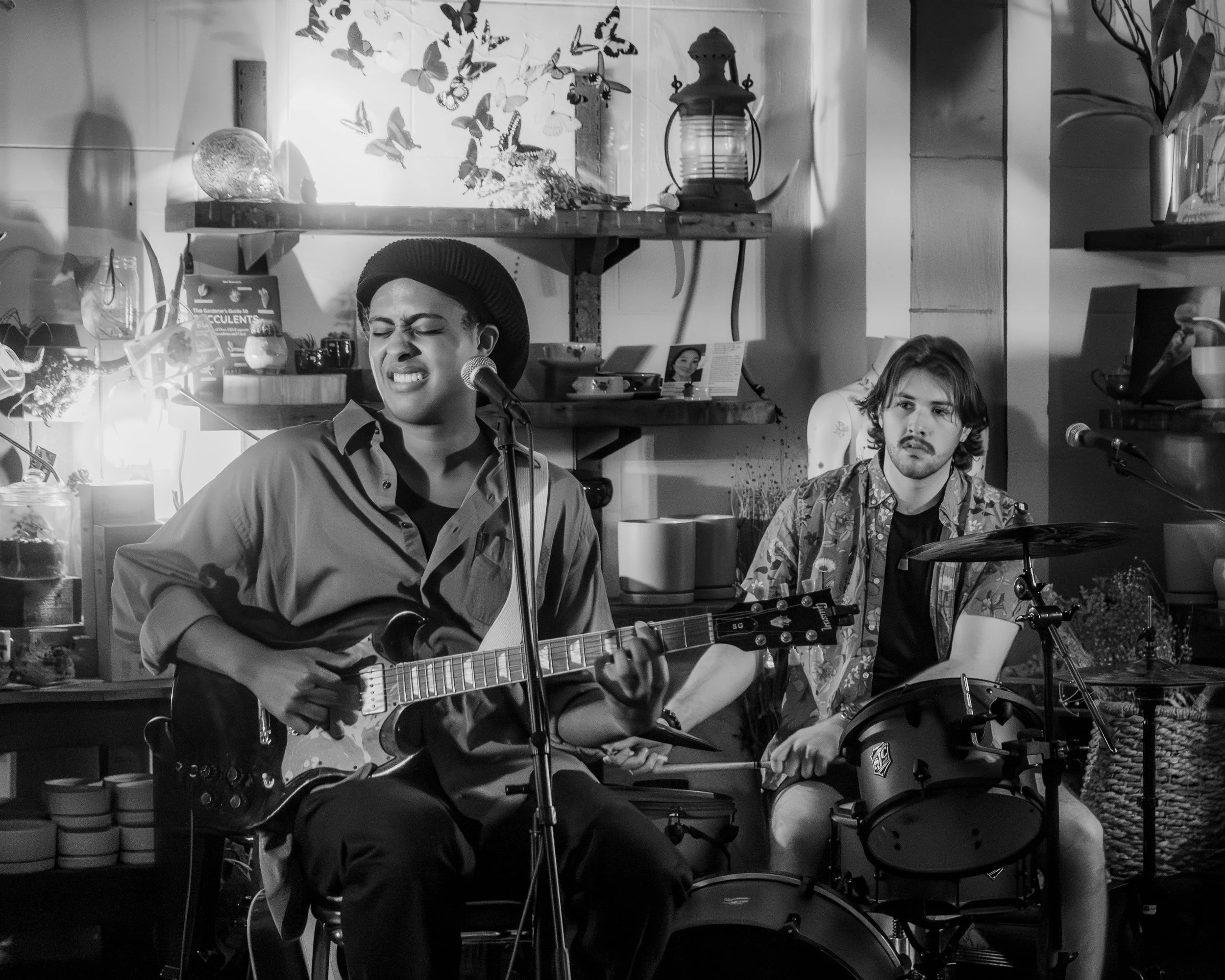 A woman singing and playing an electric guitar with a man on drums in a cozy, well-decorated room, black and white photo.