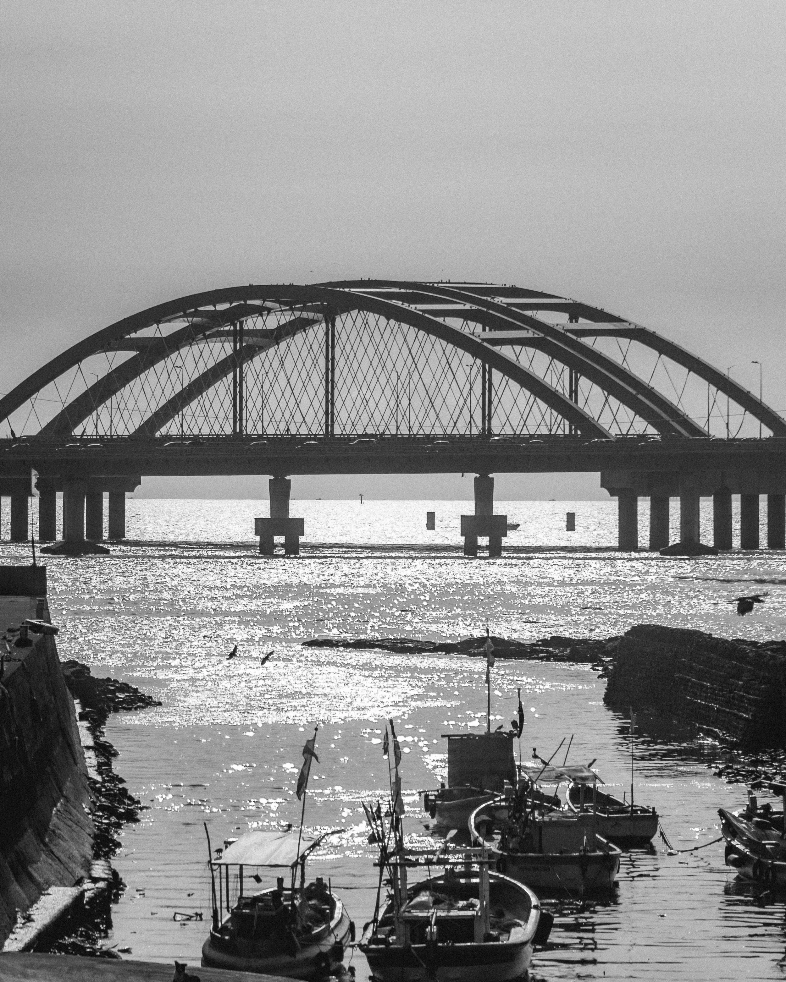 A black-and-white photograph of a large bridge over a body of water with boats in the foreground.