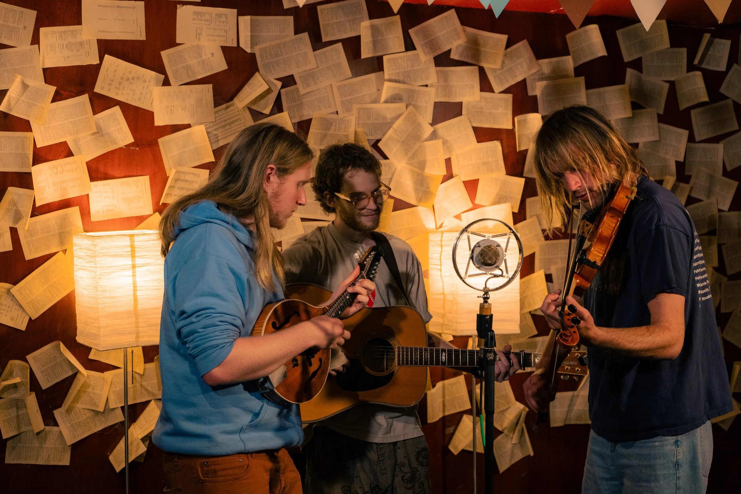 Three young men performing music in a cozy room with walls decorated with open books and paper pages, using guitars, a violin, and singing into a vintage microphone.