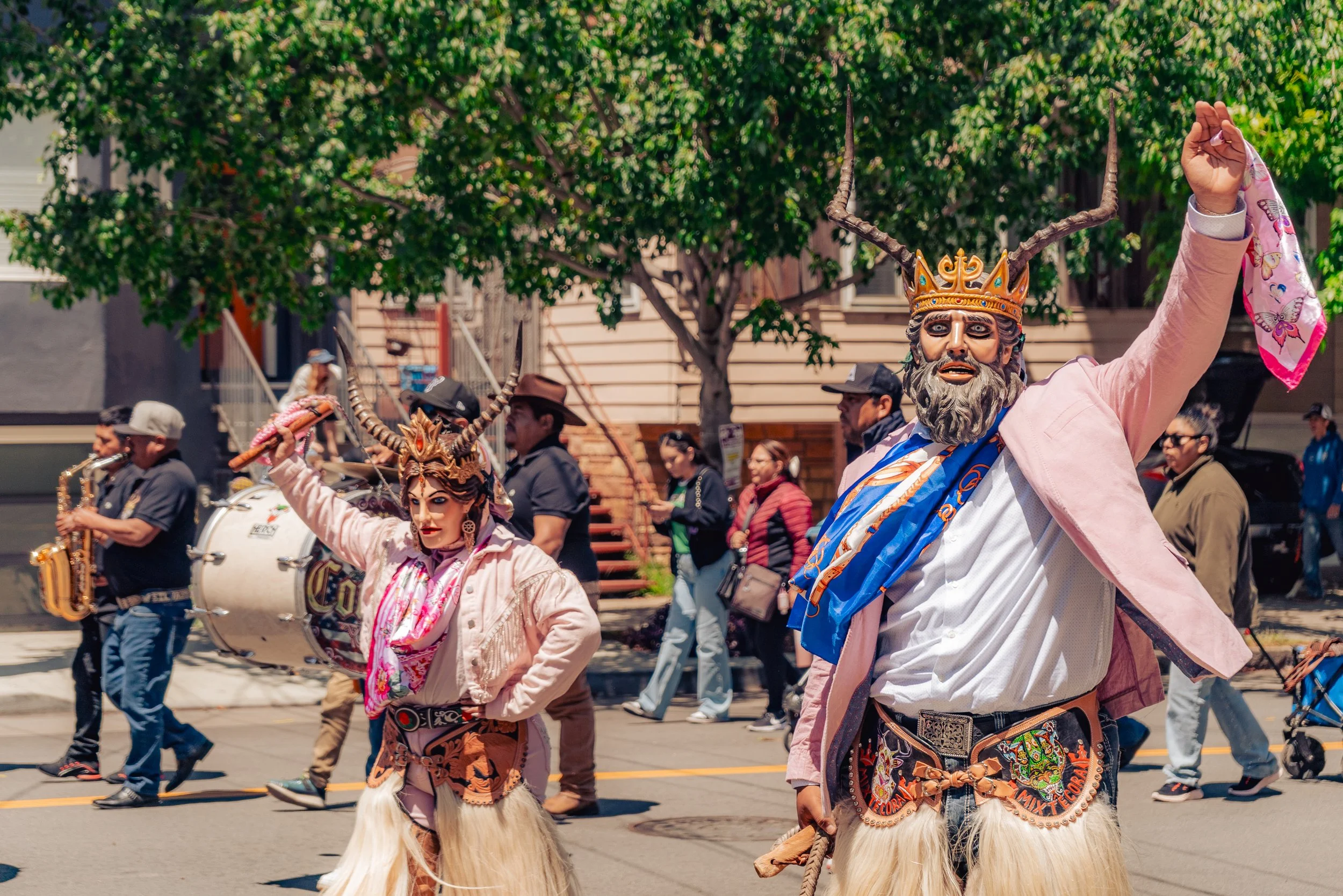 Two performers in traditional Mexican costumes with masks, horns, and elaborate accessories, participating in a parade or cultural event on a city street, with spectators and musicians in the background.