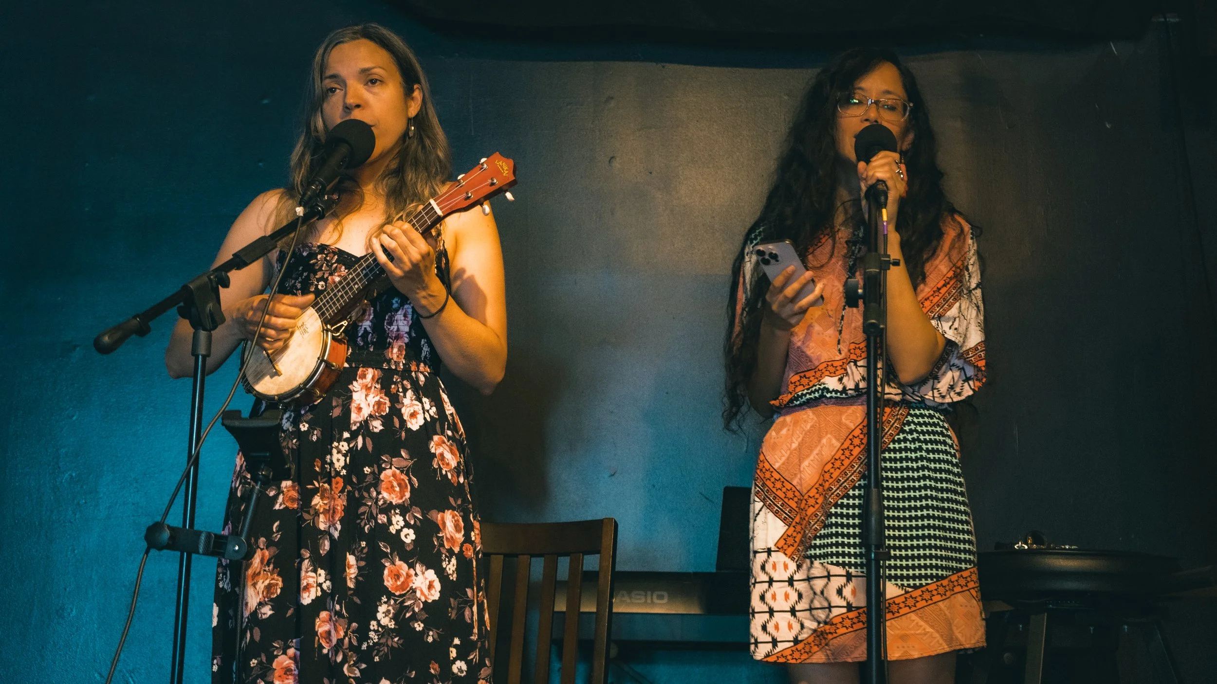 Two women standing on stage, one playing a ukulele and singing into a microphone, the other holding a phone and singing into a microphone, against a dark wall background.