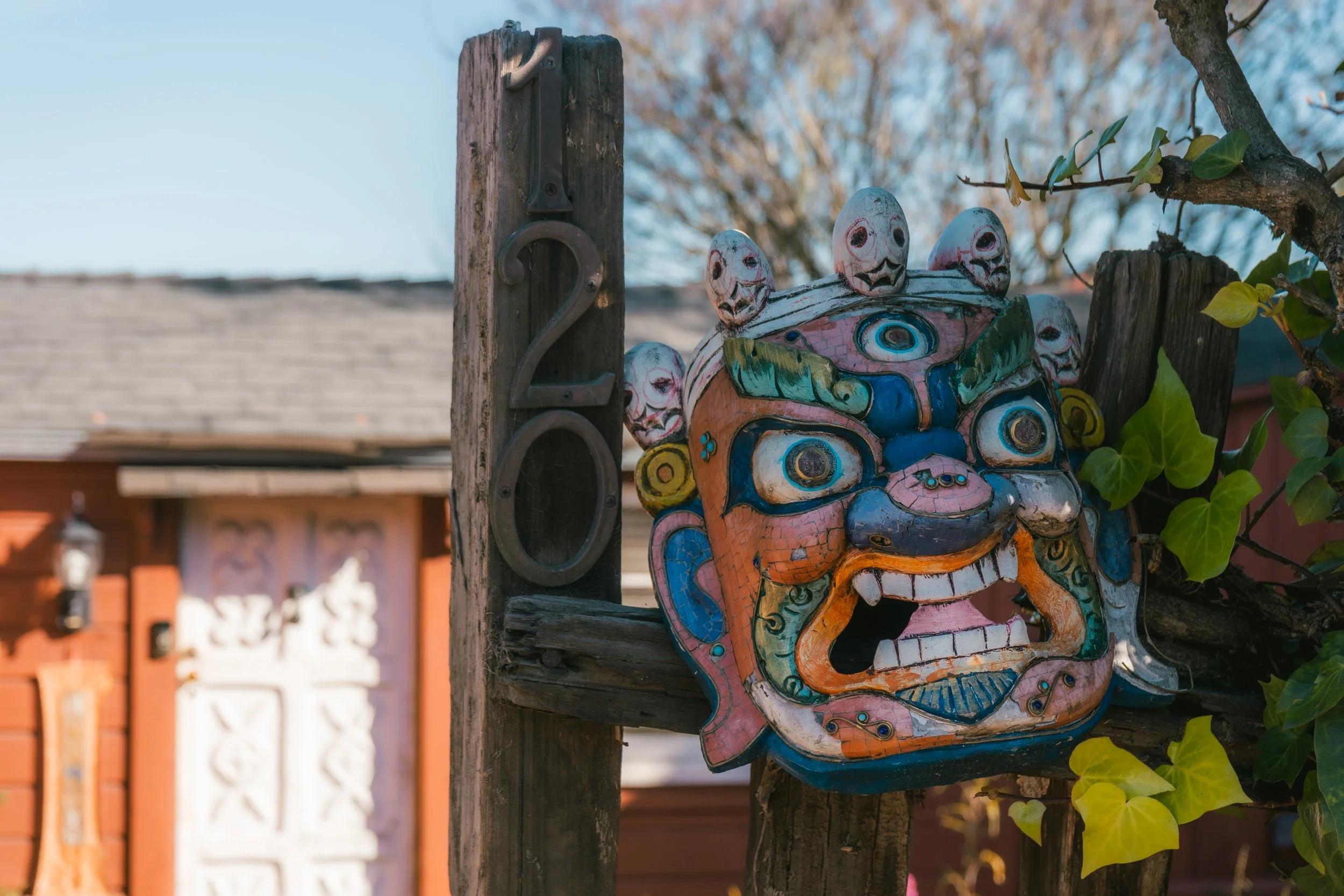 Colorful, carved, and painted face sculpture with multiple small faces on top, mounted on a wooden post outdoors with a background of a building and trees.