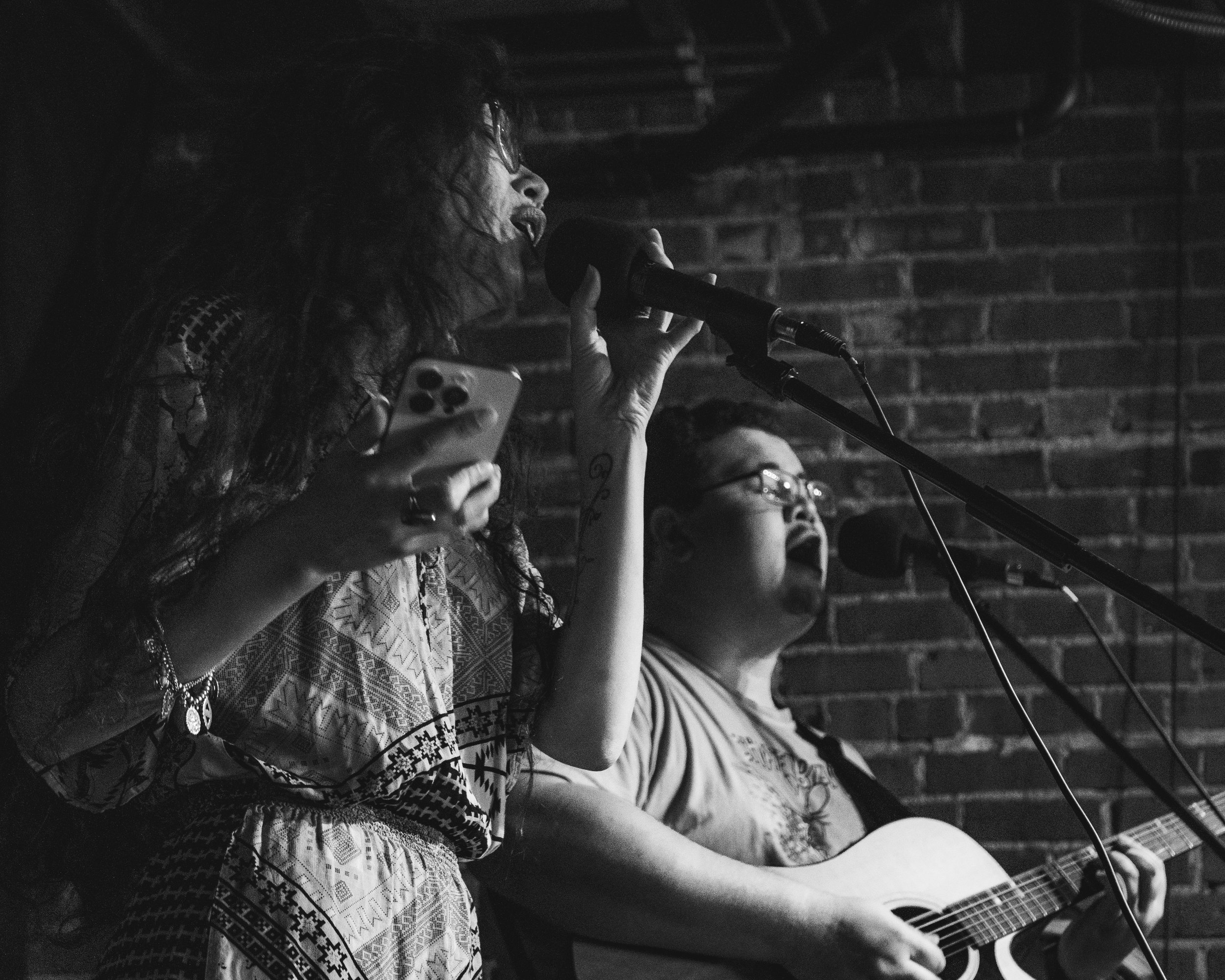 Two musicians perform live on stage against a brick wall. The female singer on the left is singing into a microphone and holding a smartphone. The male guitarist on the right is singing into a microphone and playing an acoustic guitar.
