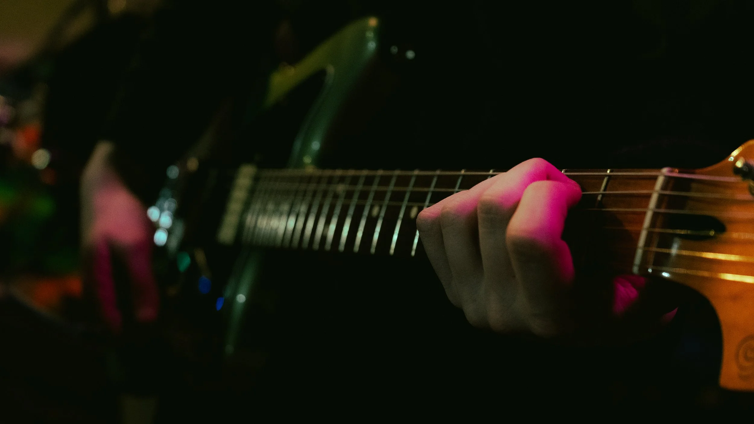 Close-up of a person's hand pressing guitar strings on the fretboard in a dimly lit environment with pink and green lighting.