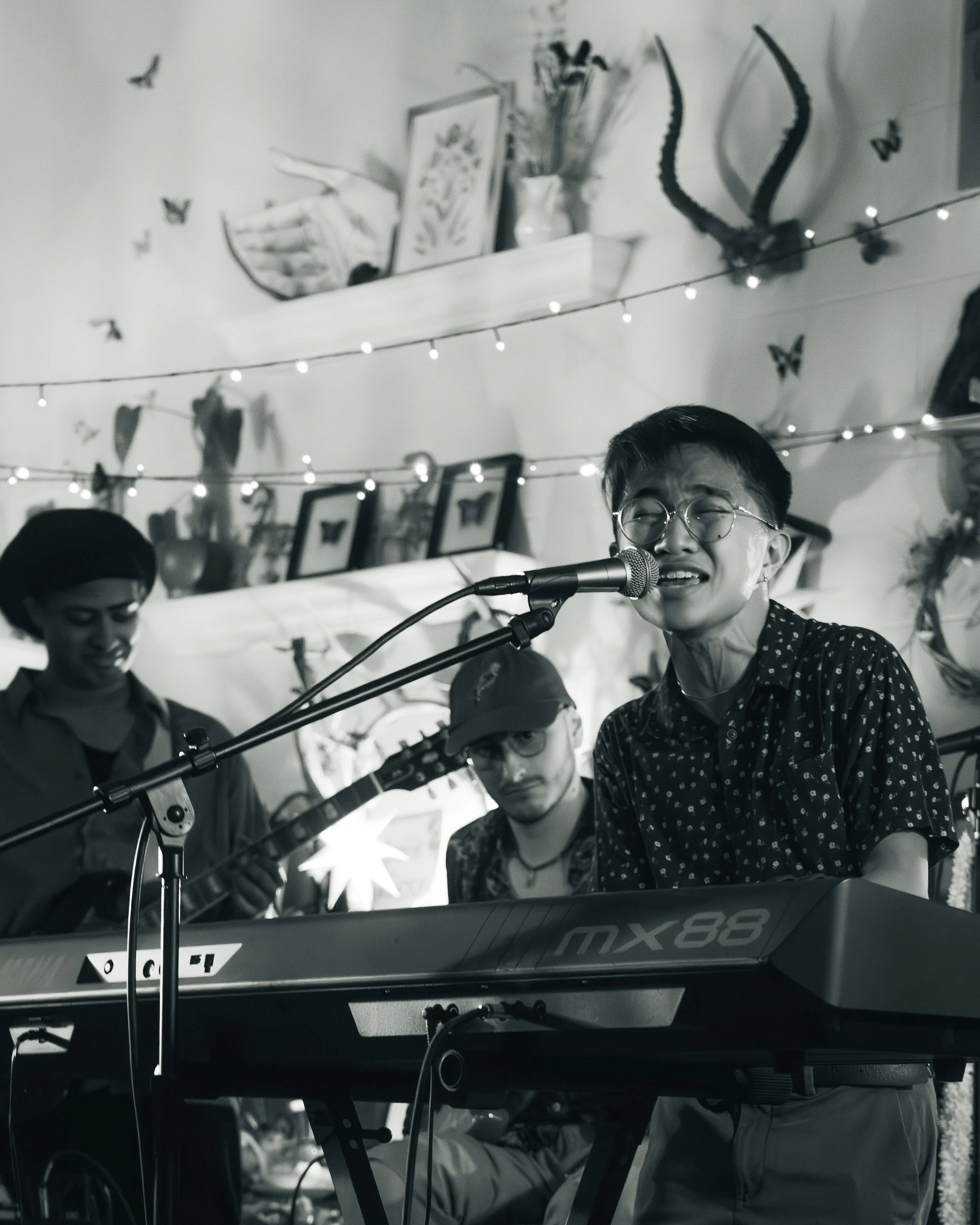 A person singing and playing a keyboard, with two other people in the background, in a decorated indoor setting with hanging string lights, framed pictures, and wall art.