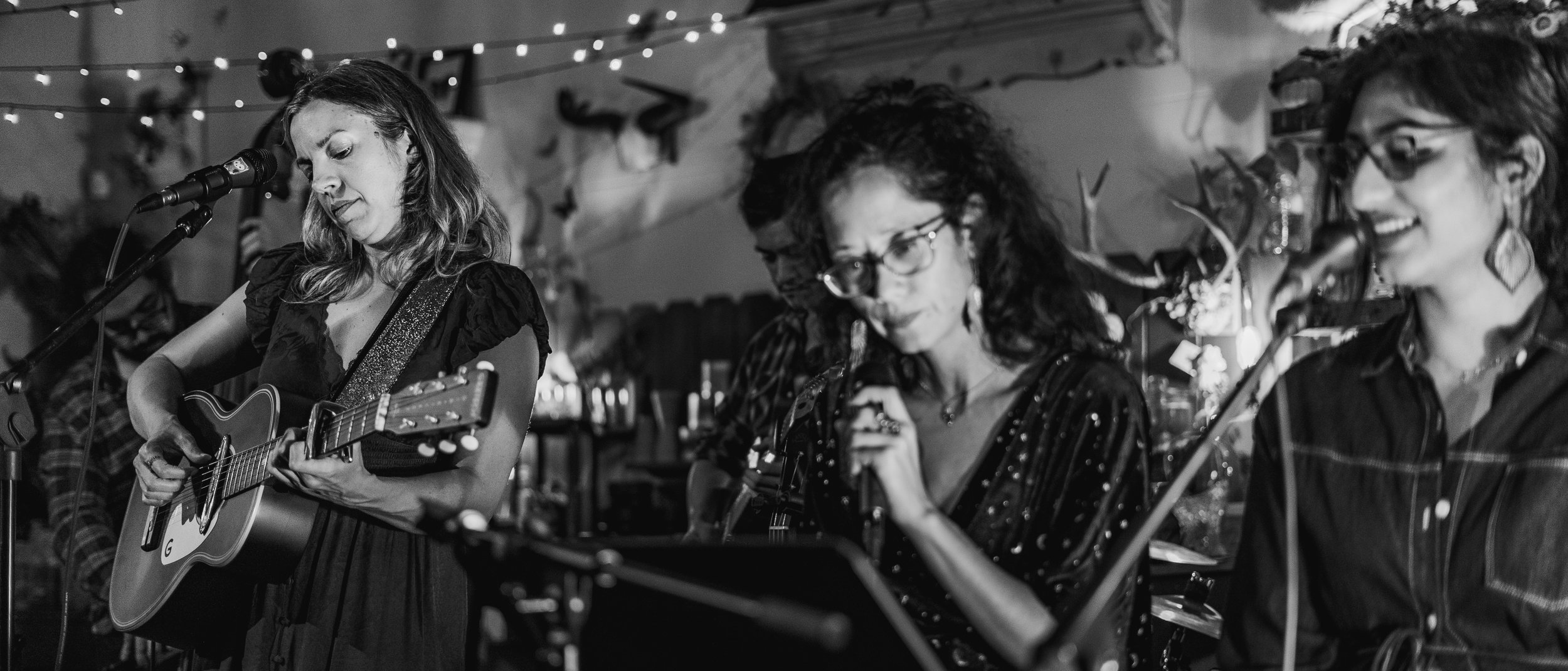 Four women performing music together; one playing guitar and singing into a microphone, two others singing into microphones, all wearing glasses and casual attire, in a cozy, decorated indoor setting.