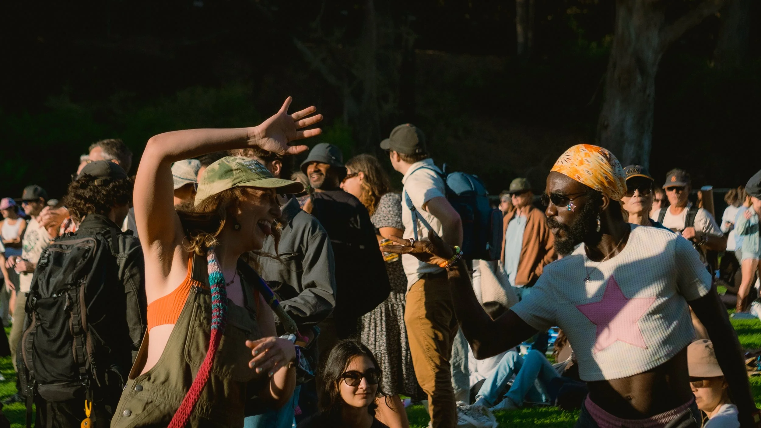People socializing outdoors at a gathering with trees in the background, some standing, some sitting, in a park or festival setting.