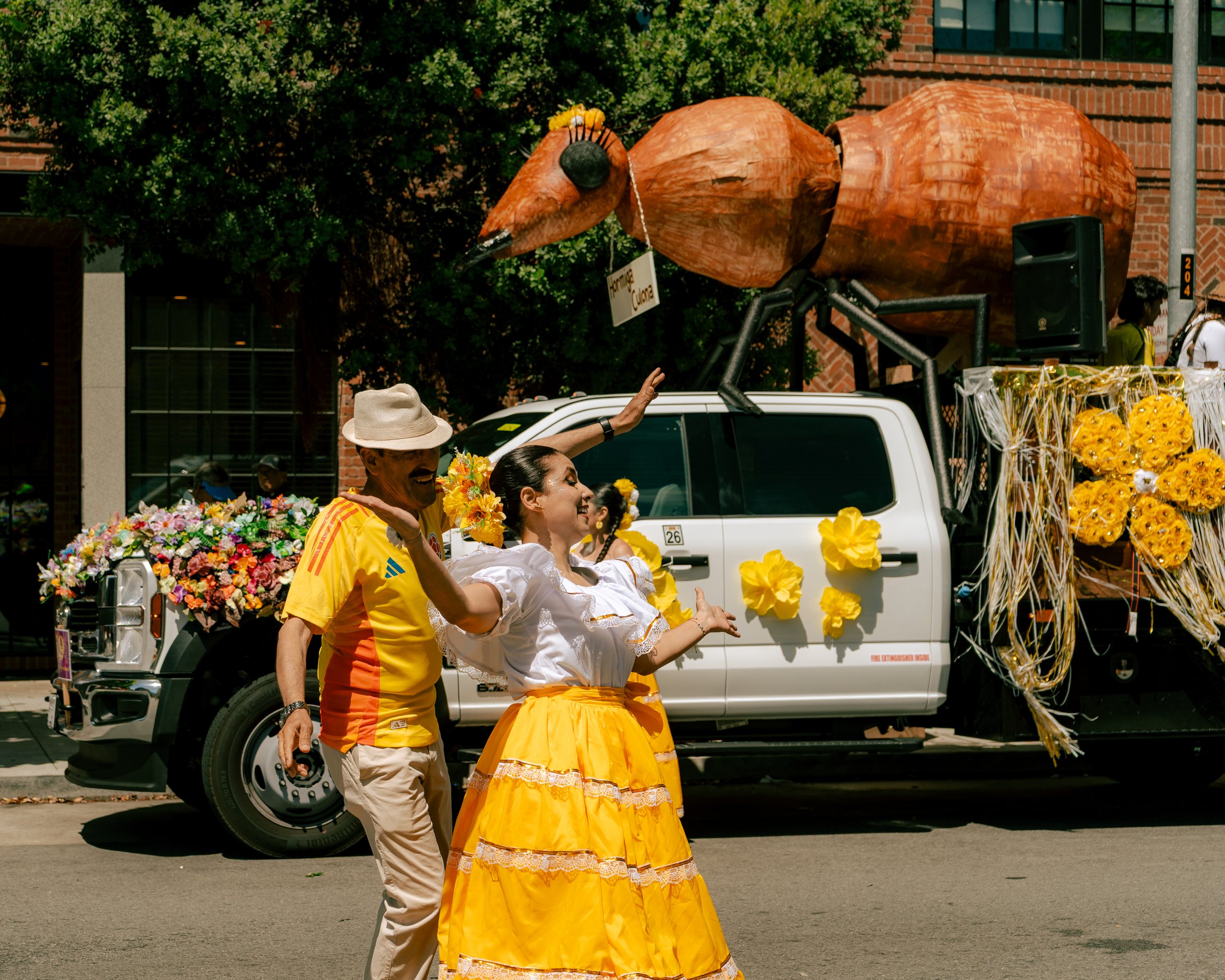 People dancing in front of a decorated parade float with a giant ant sculpture, yellow floral decorations, and colorful flowers, during a parade celebration.