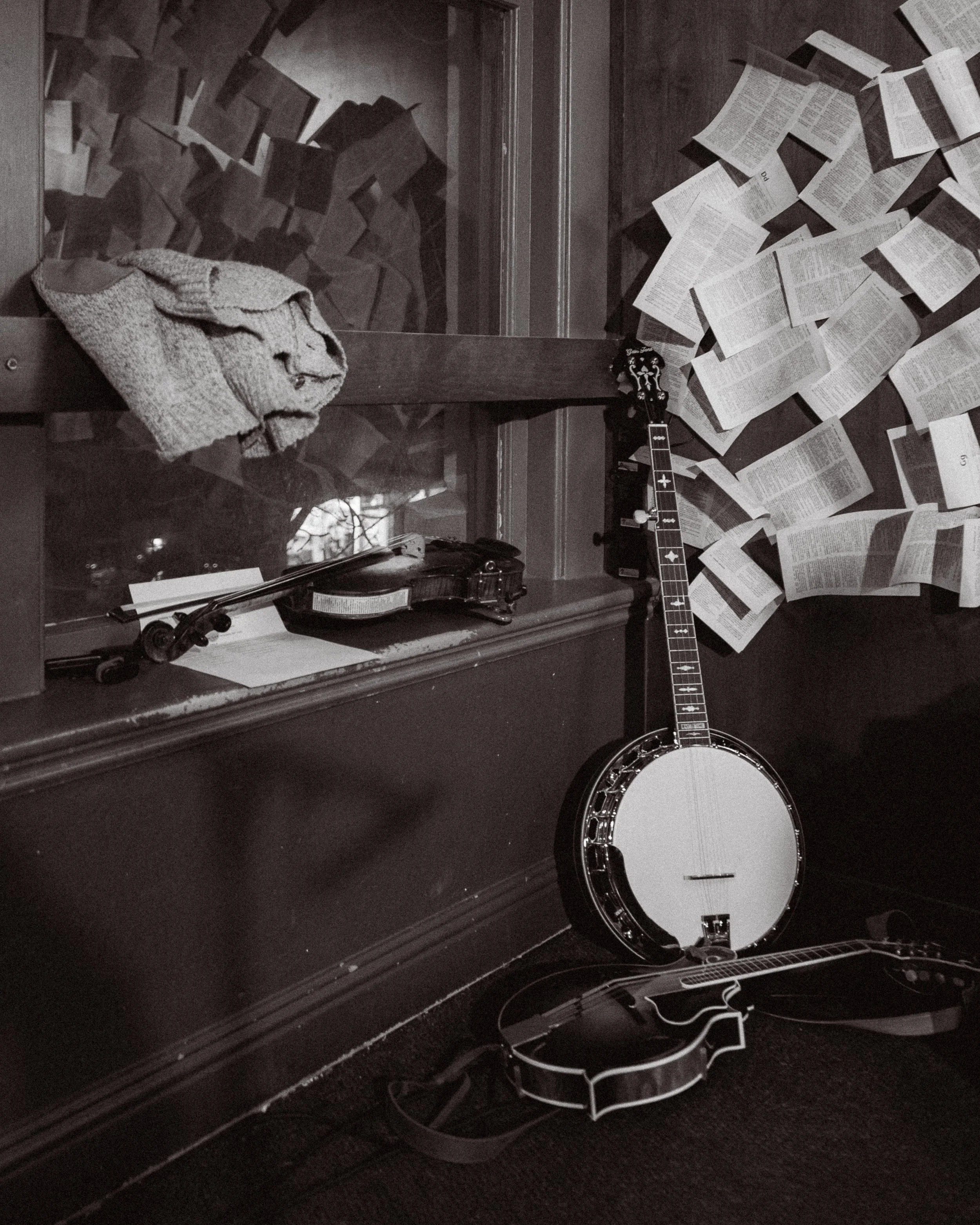 A black and white photo of a windowsill with a folded knitted hat, a violin, a banjo, and sheet music scattered around, with a wall covered in pages of printed text.
