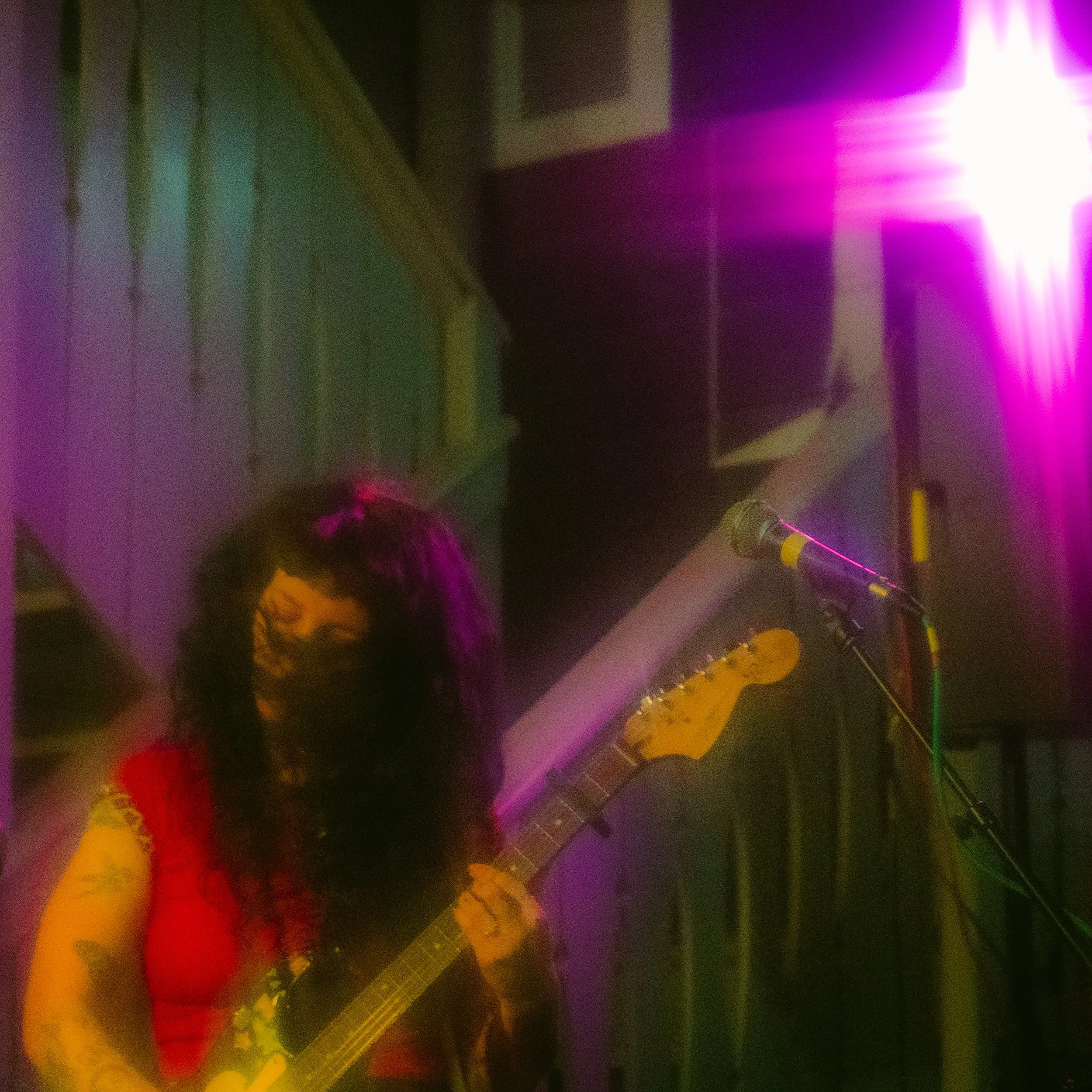 A woman with long dark hair playing an electric guitar on stage under purple and pink stage lights, with a microphone nearby.