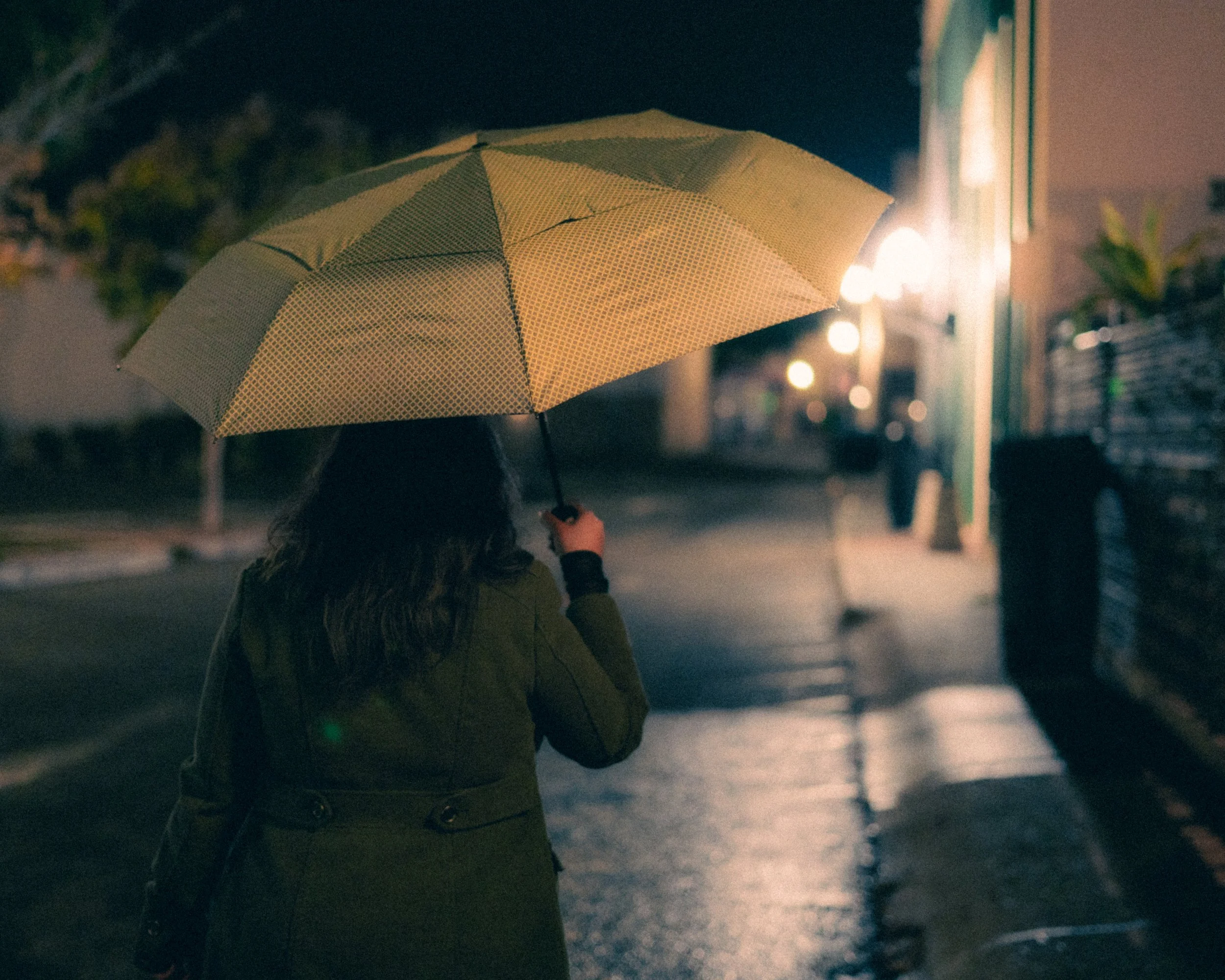 A person with dark hair walking on a wet sidewalk at night, holding a yellow umbrella, illuminated by distant streetlights and shop lights.