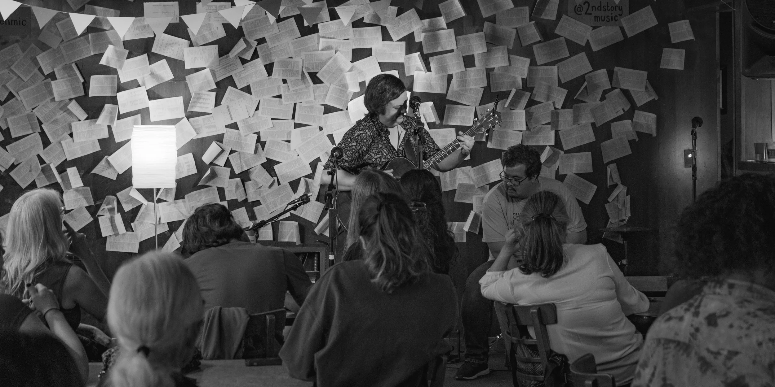 A woman playing an acoustic guitar and singing on a small stage, with an audience watching, in a cozy venue with a wall decorated with numerous open books or sheets of paper.