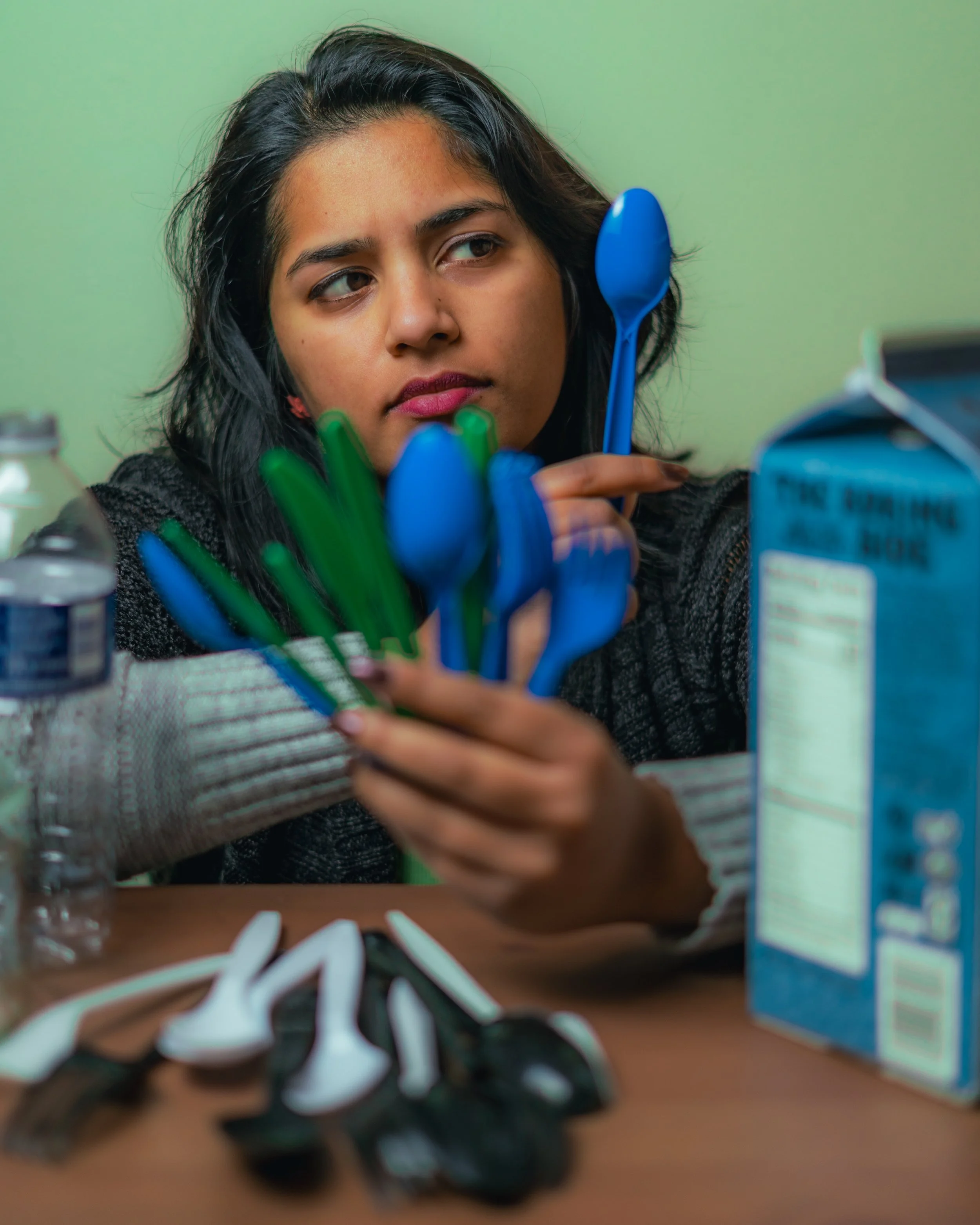 A young woman with dark, wavy hair holds blue and green plastic spoons and utensils, sitting at a table with a box of cereal, a jar, and a bunch of used black and white plastic spoons in front of her. She appears to be in a kitchen or dining area wit