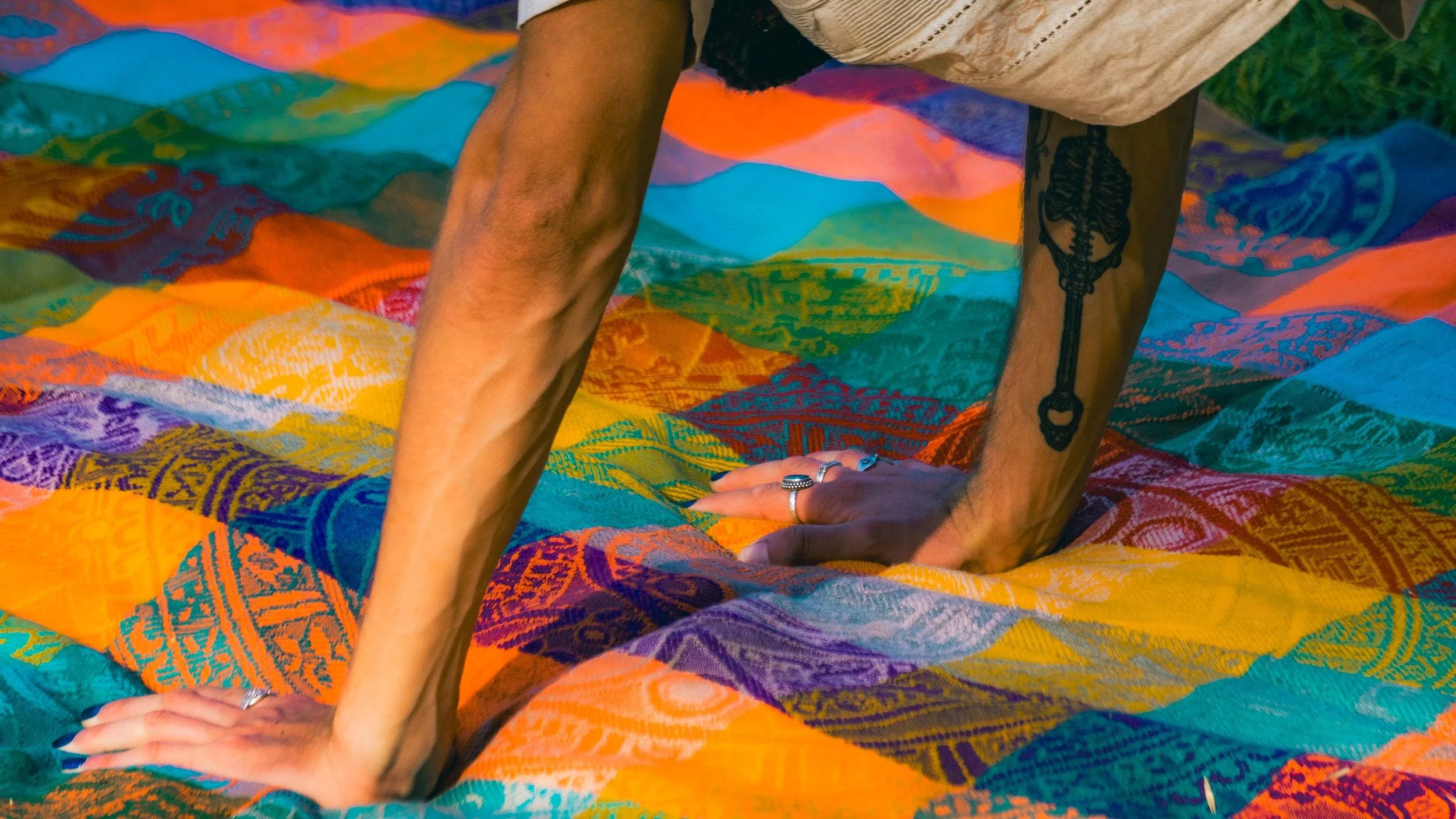 Person practicing yoga on colorful patterned blanket, with visible tattoo of a key on their arm, and rings on their fingers.